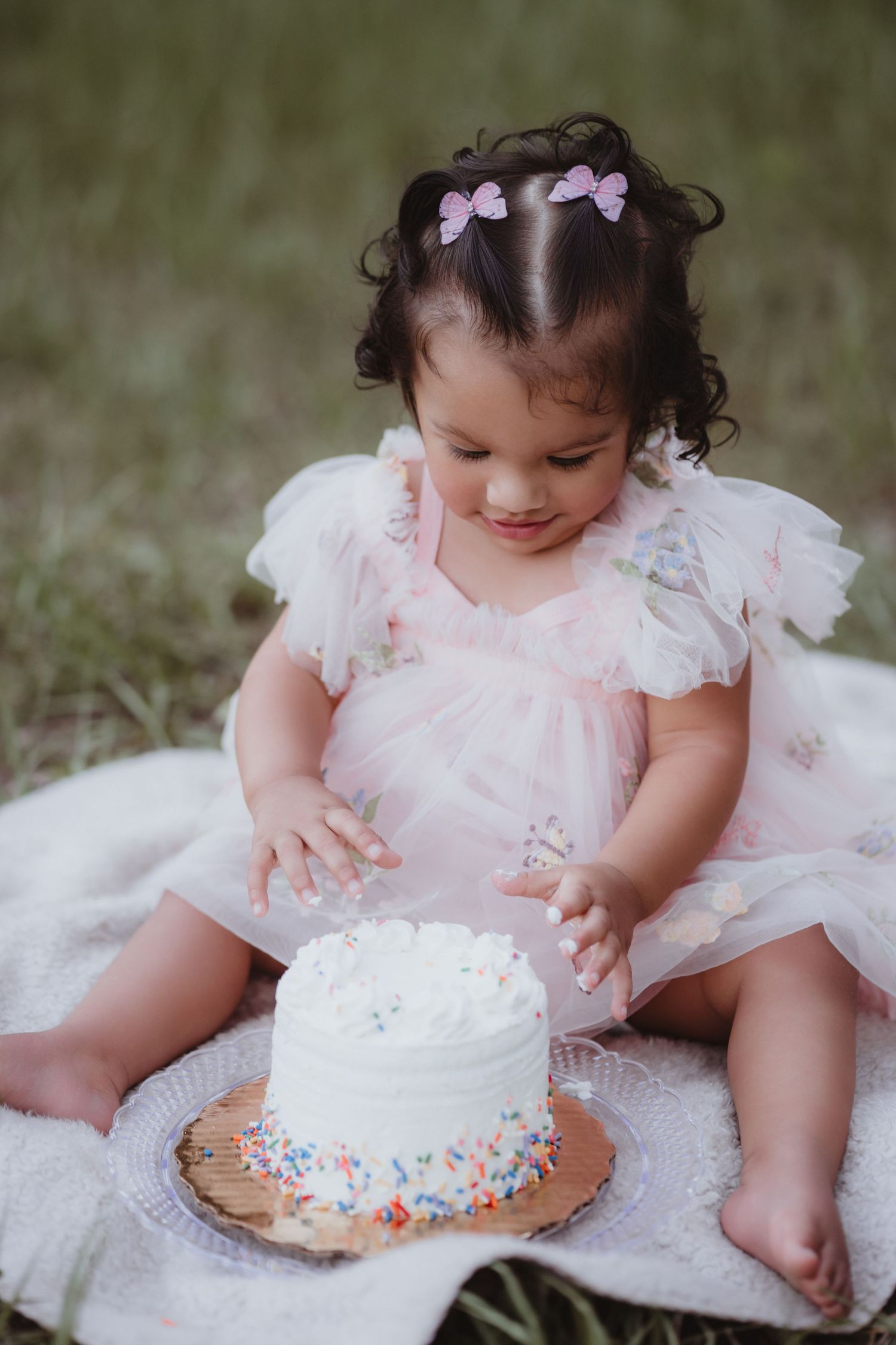 Baby girl in pink tulle dress touching white sprinkle smash cake outdoors.