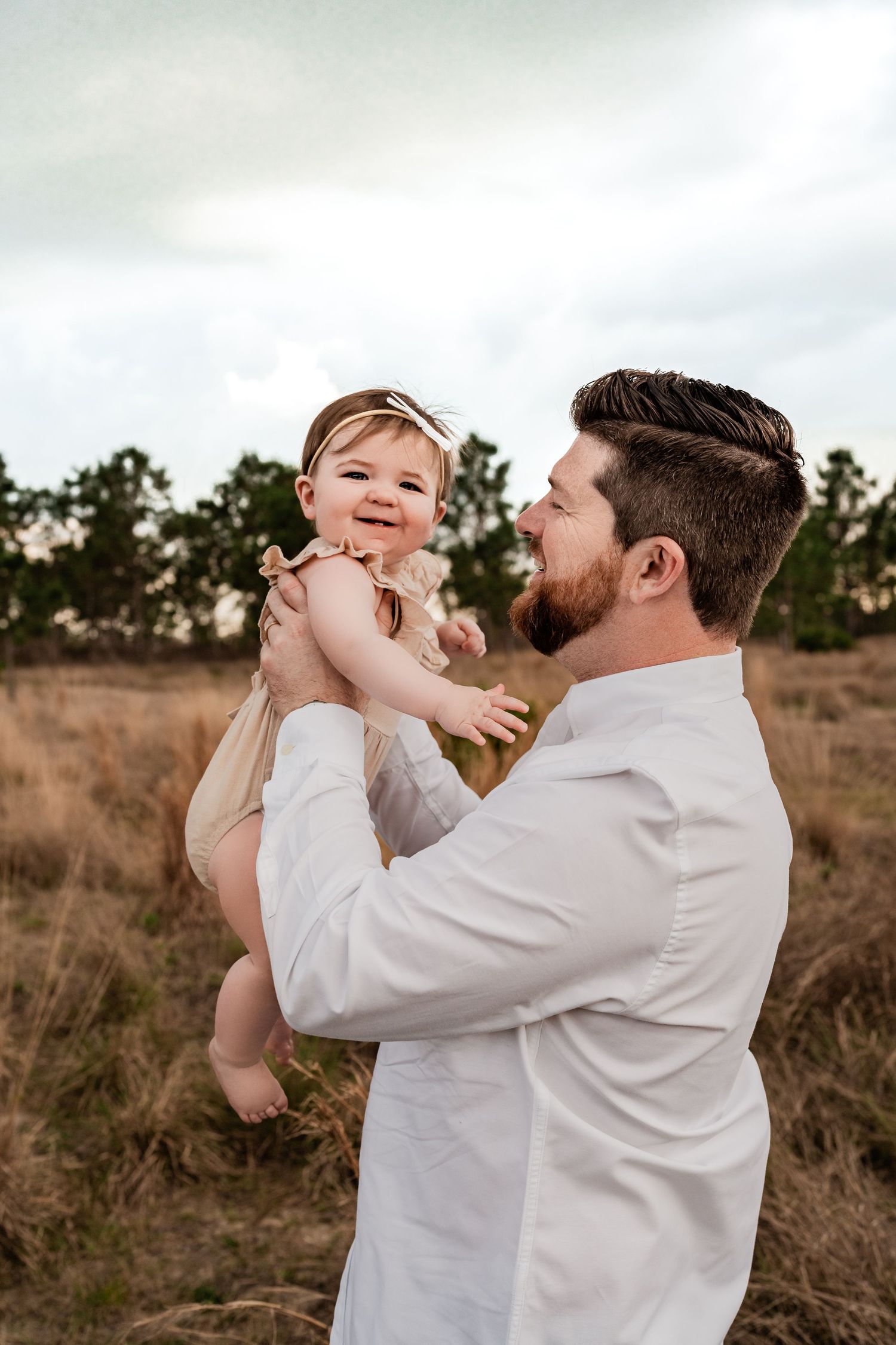 Happy baby girl in tan romper laughing as father lifts her in a golden grass field.