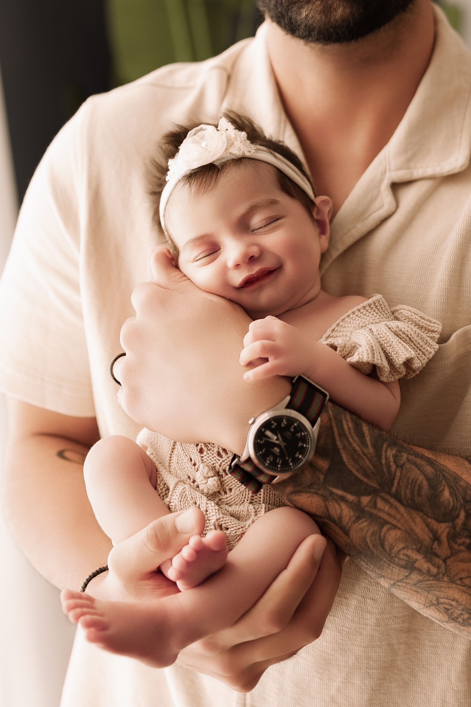 Bearded tattooed father cradling smiling newborn baby girl in floral headband and knit romper.