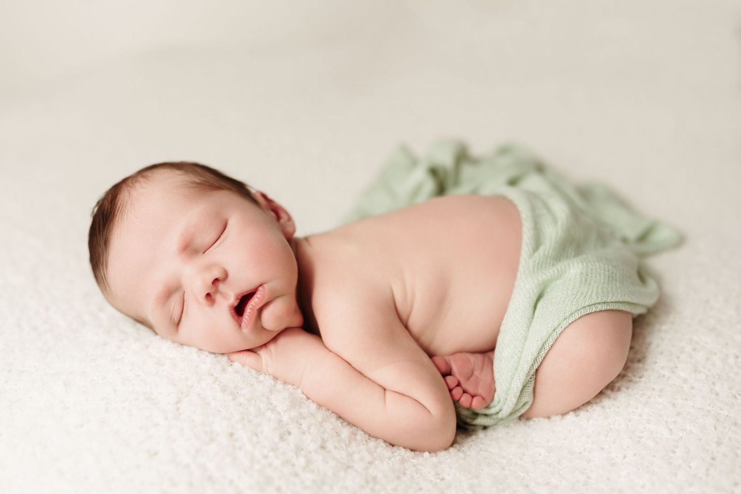Peaceful newborn baby sleeping on white fur wrapped in soft green knit blanket.