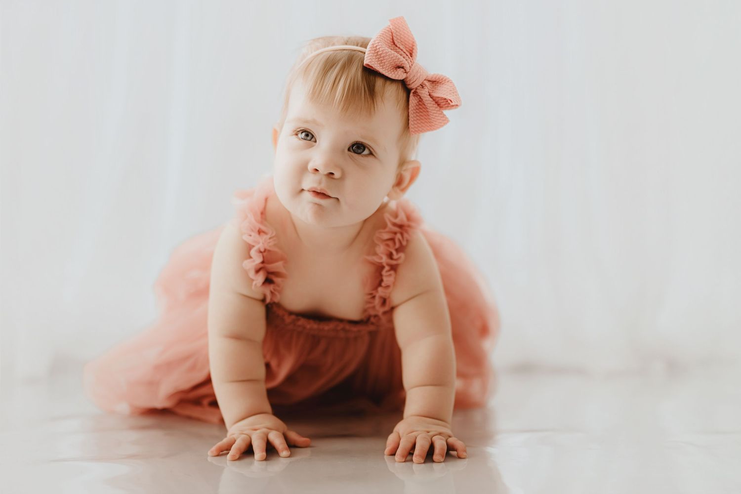 Adorable baby girl in pink tulle dress and bow headband crawling on white floor.