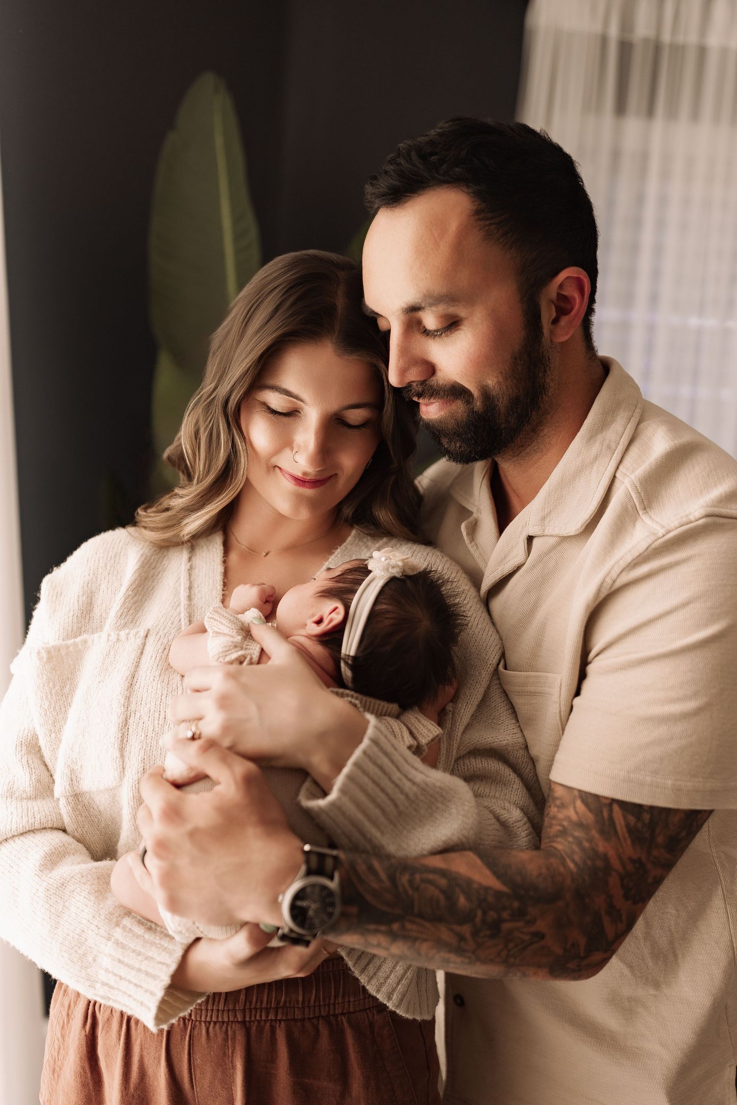 Loving parents in neutral tones tenderly gazing at their sleeping newborn baby with bow headband.