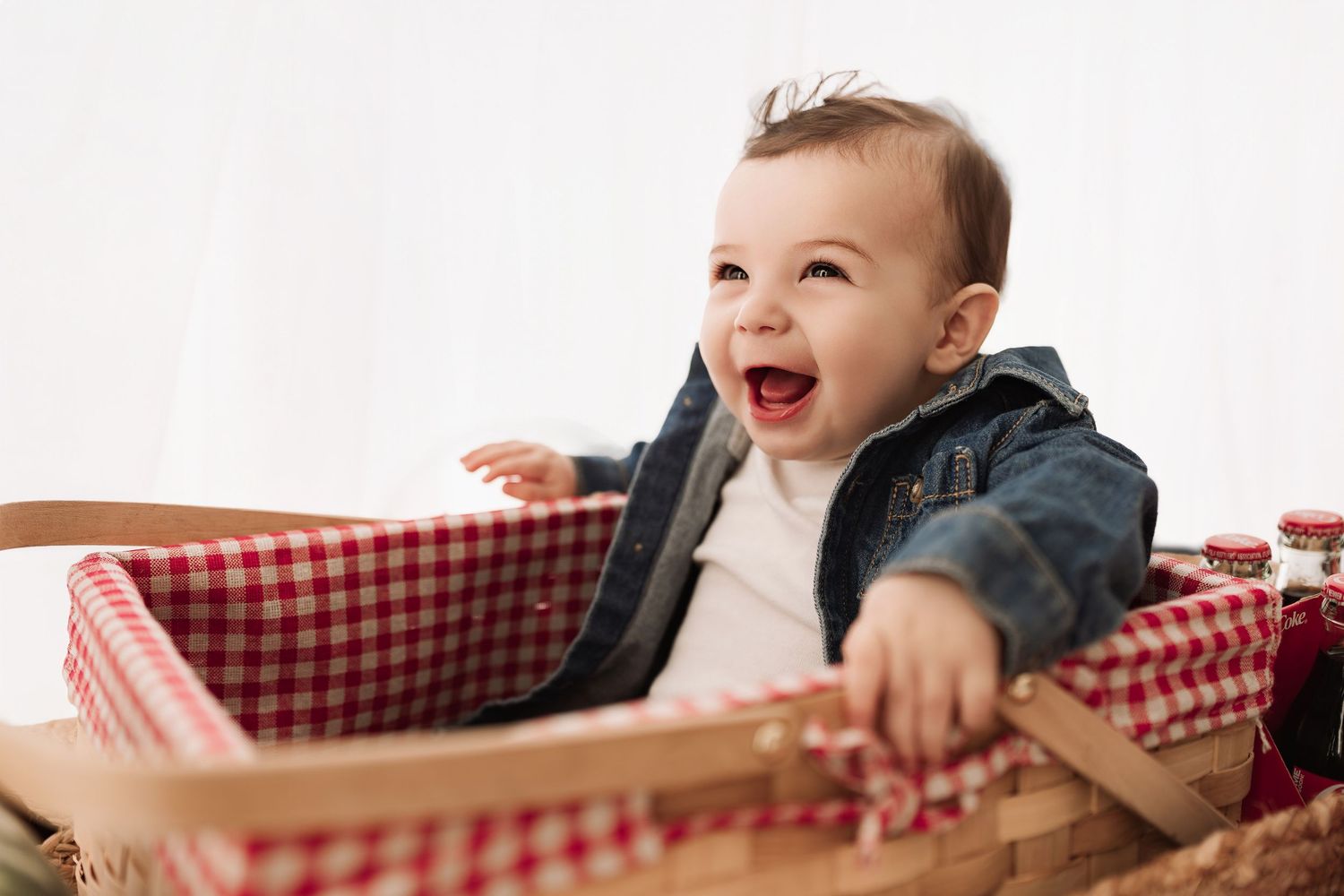 Cheerful baby in denim jacket laughing with mouth wide open while sitting in a picnic basket.