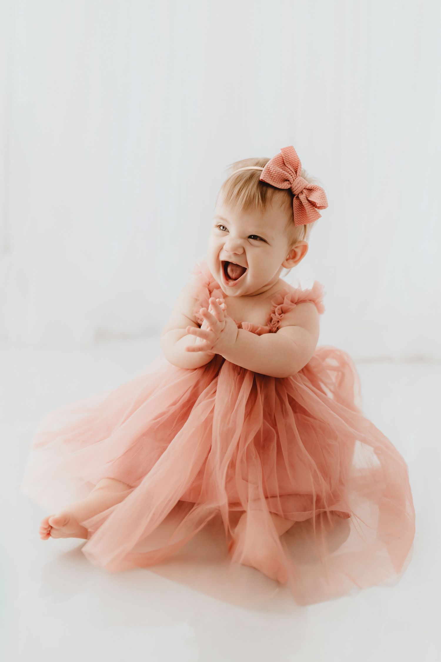 Joyful baby girl laughing in a flowing pink tulle dress with matching bow headband.