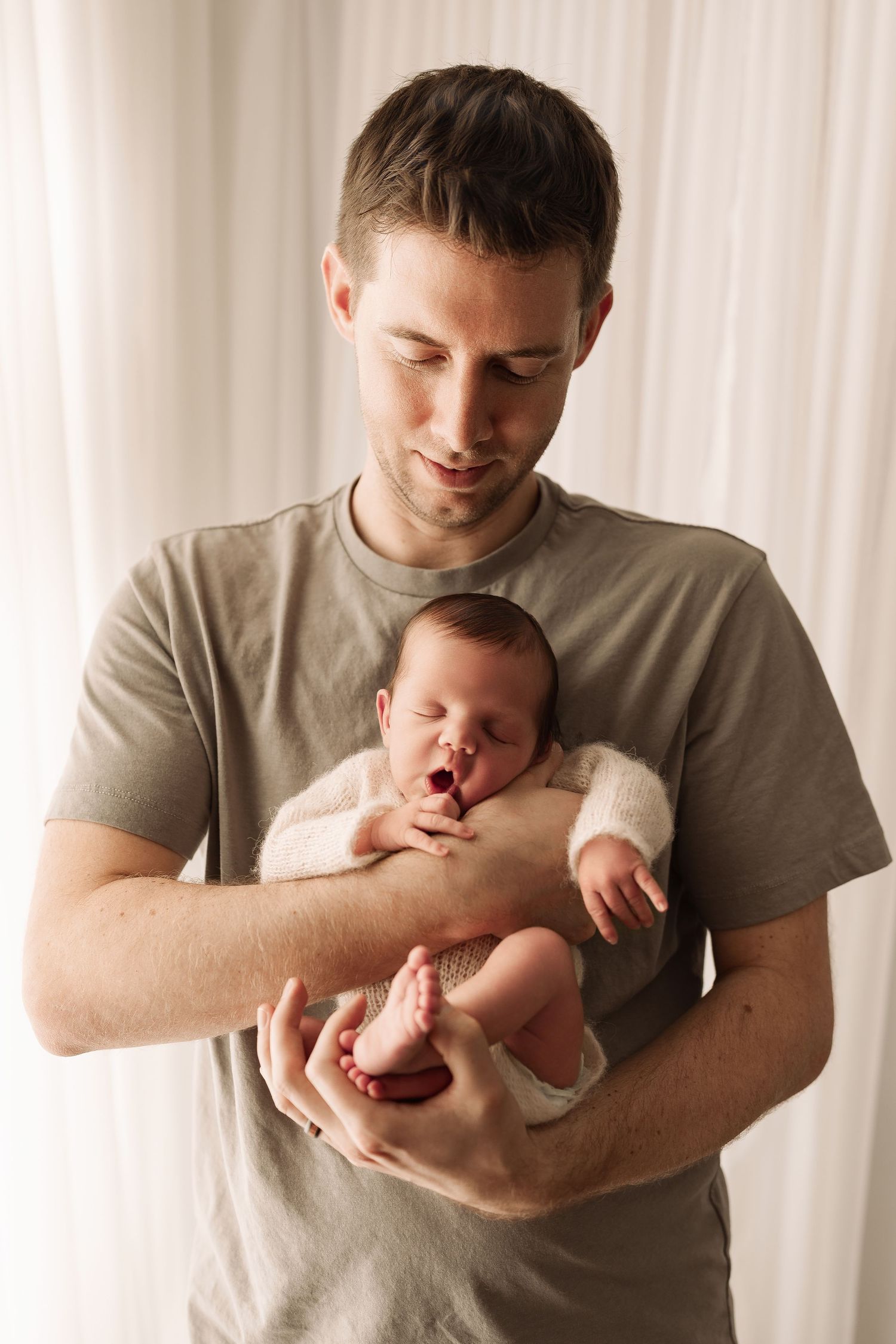Young father in gray t-shirt holding his yawning newborn baby against a white curtain backdrop.