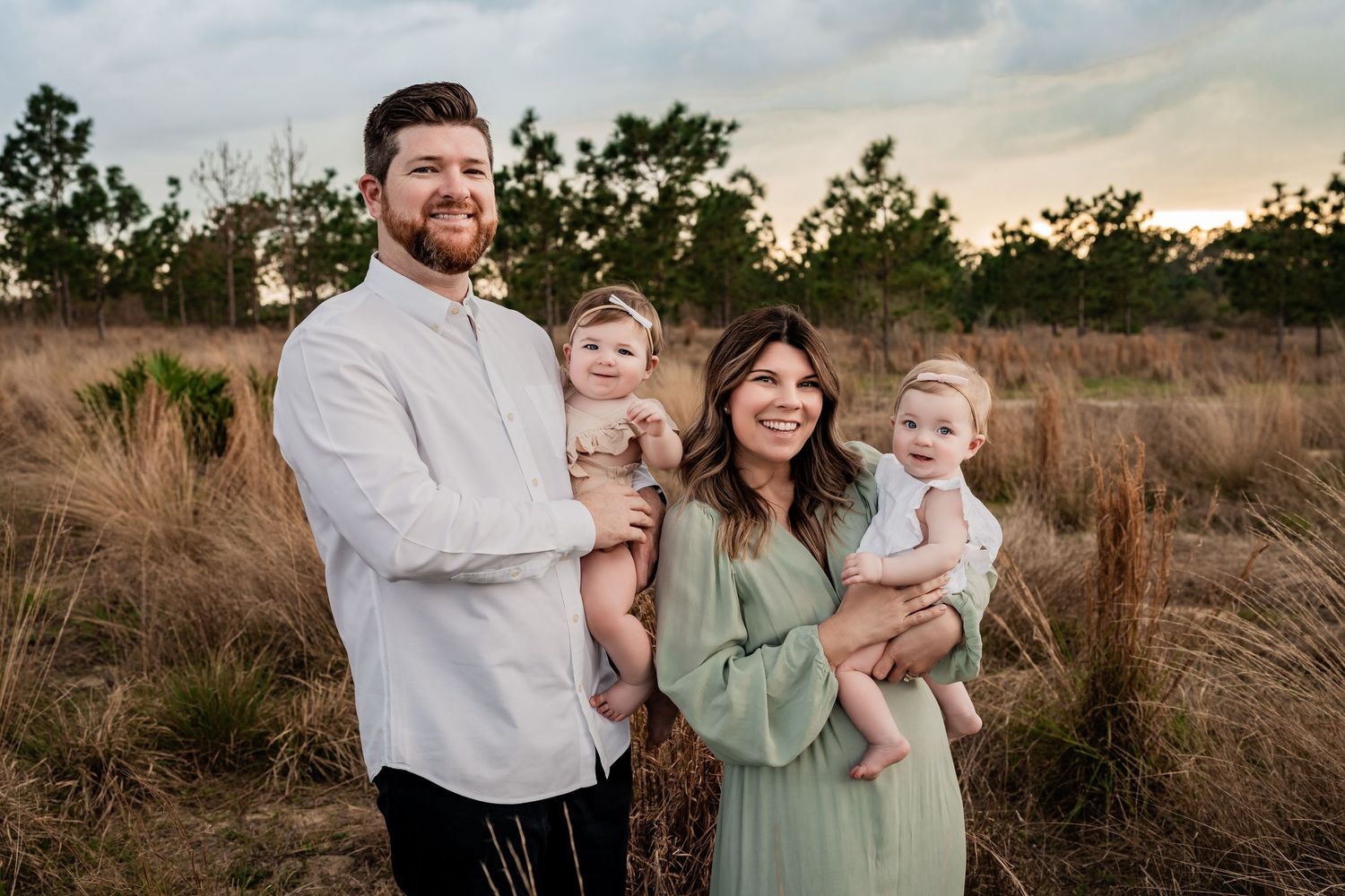 Family of four smiling outdoors in golden grass field at sunset, each parent holding a baby.