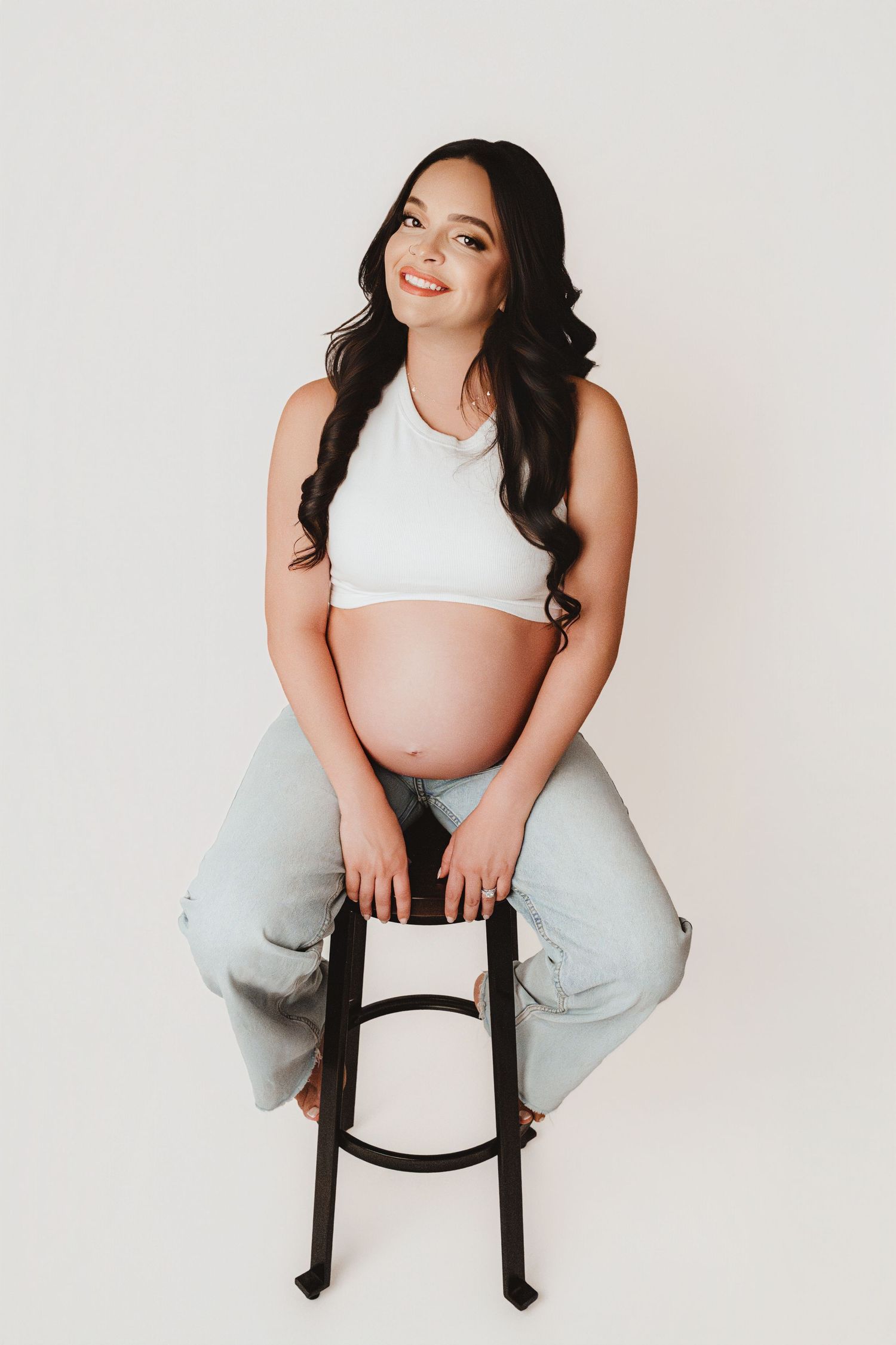 Smiling pregnant woman with long dark hair sitting on stool in white crop top and jeans.