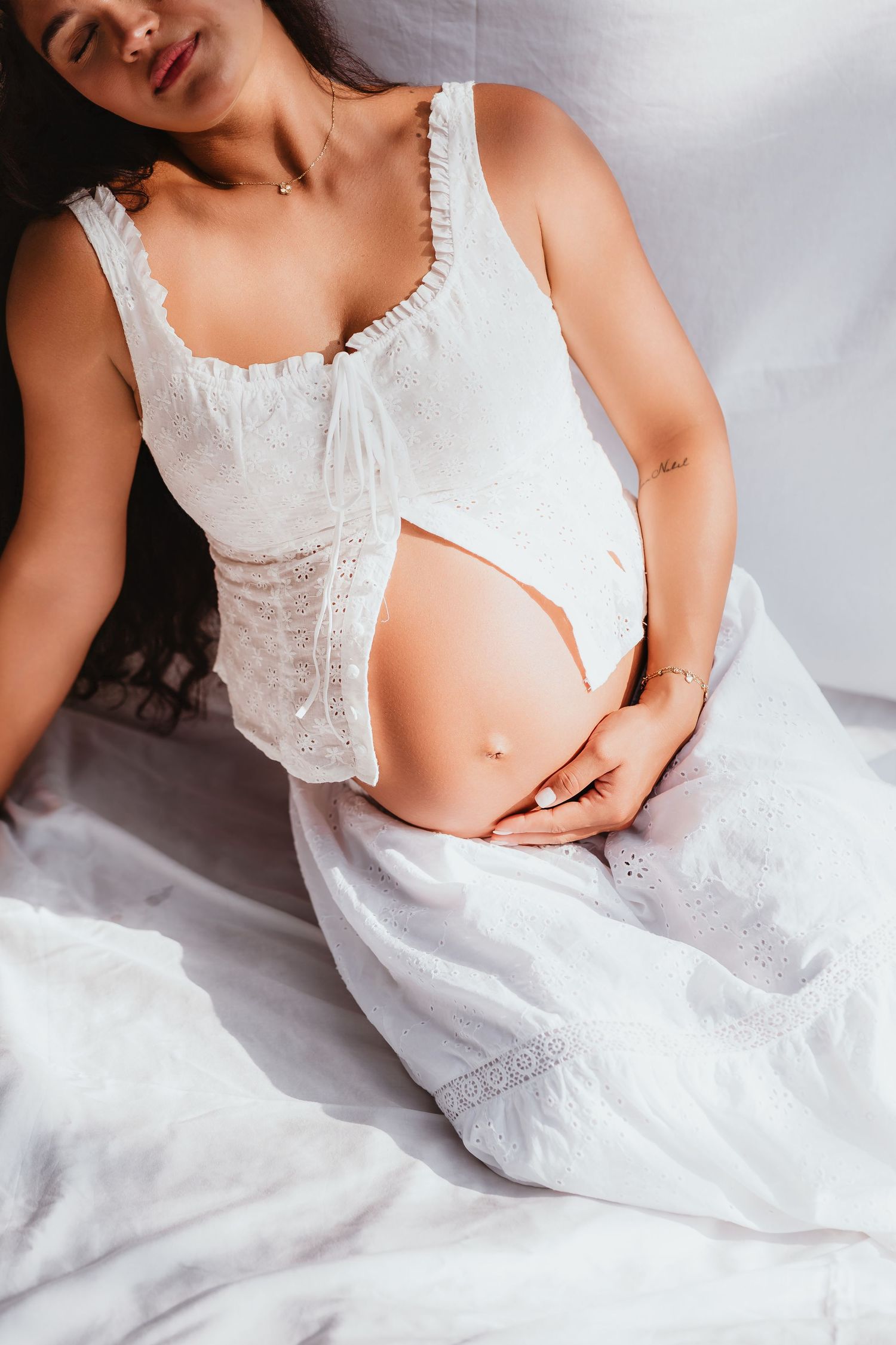Close-up of pregnant woman in white lace dress gently holding bare baby bump while seated.
