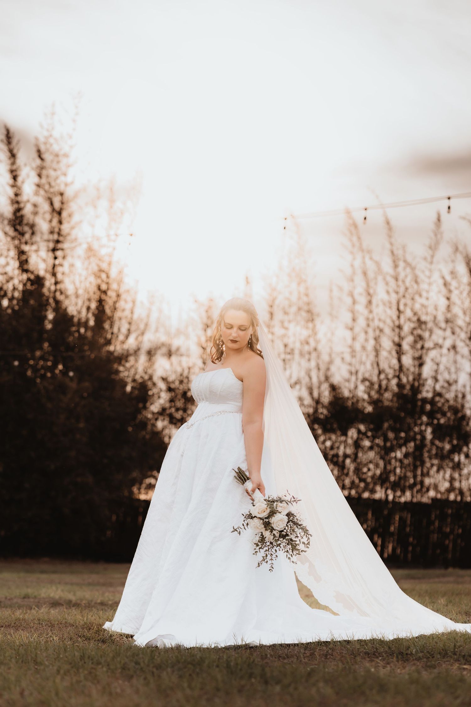 Bride in strapless white ball gown with long veil holds bouquet at sunset outdoors.