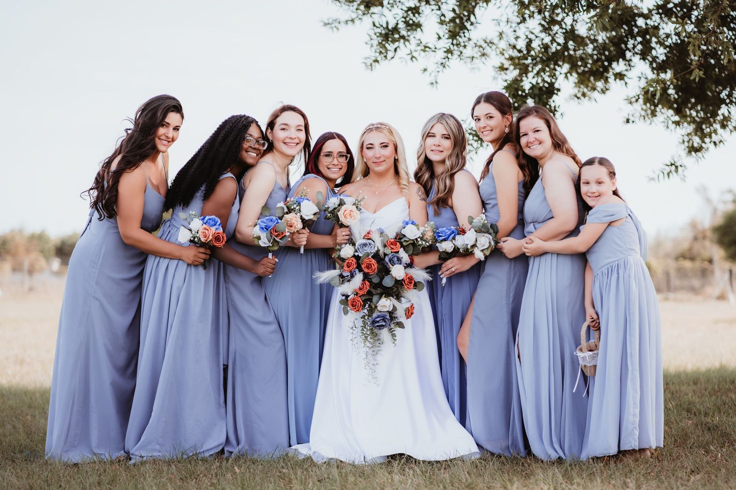 Bride in white gown with seven bridesmaids in lavender dresses holding colorful bouquets outdoors.