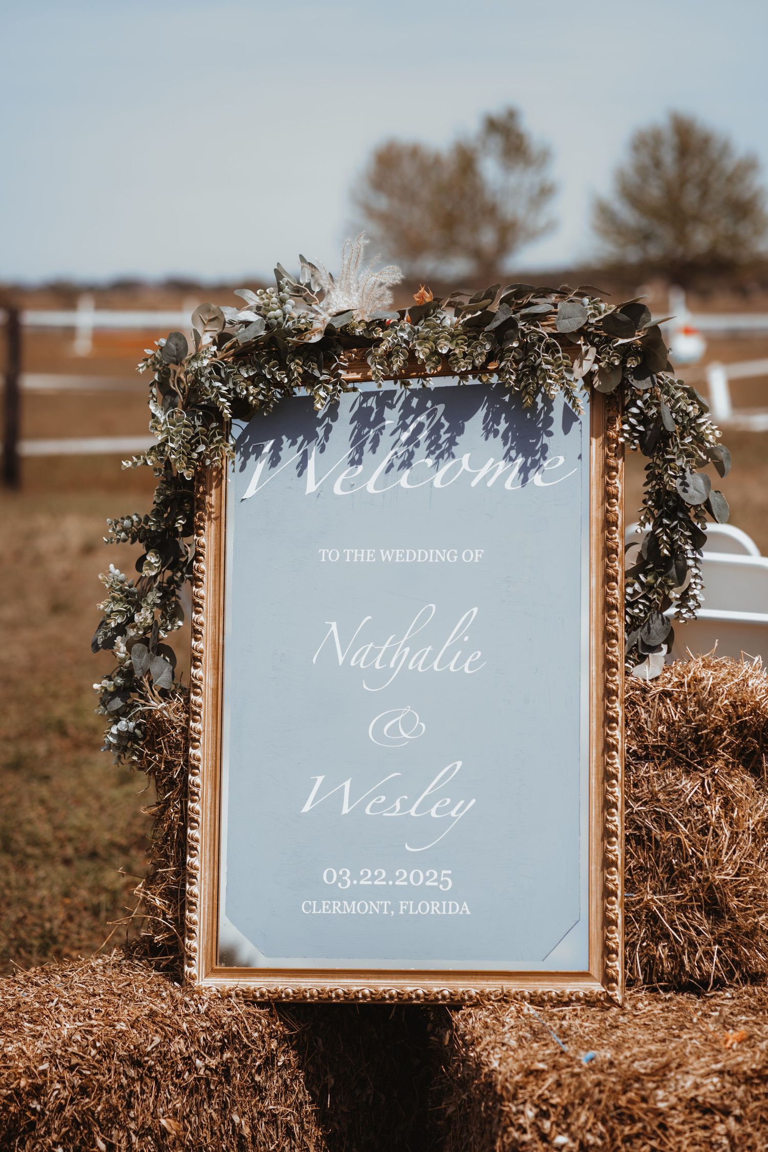 Elegant gold-framed wedding welcome mirror sign for Nathalie and Wesley dated 03.22.2025 in Clermont Florida on hay bale.