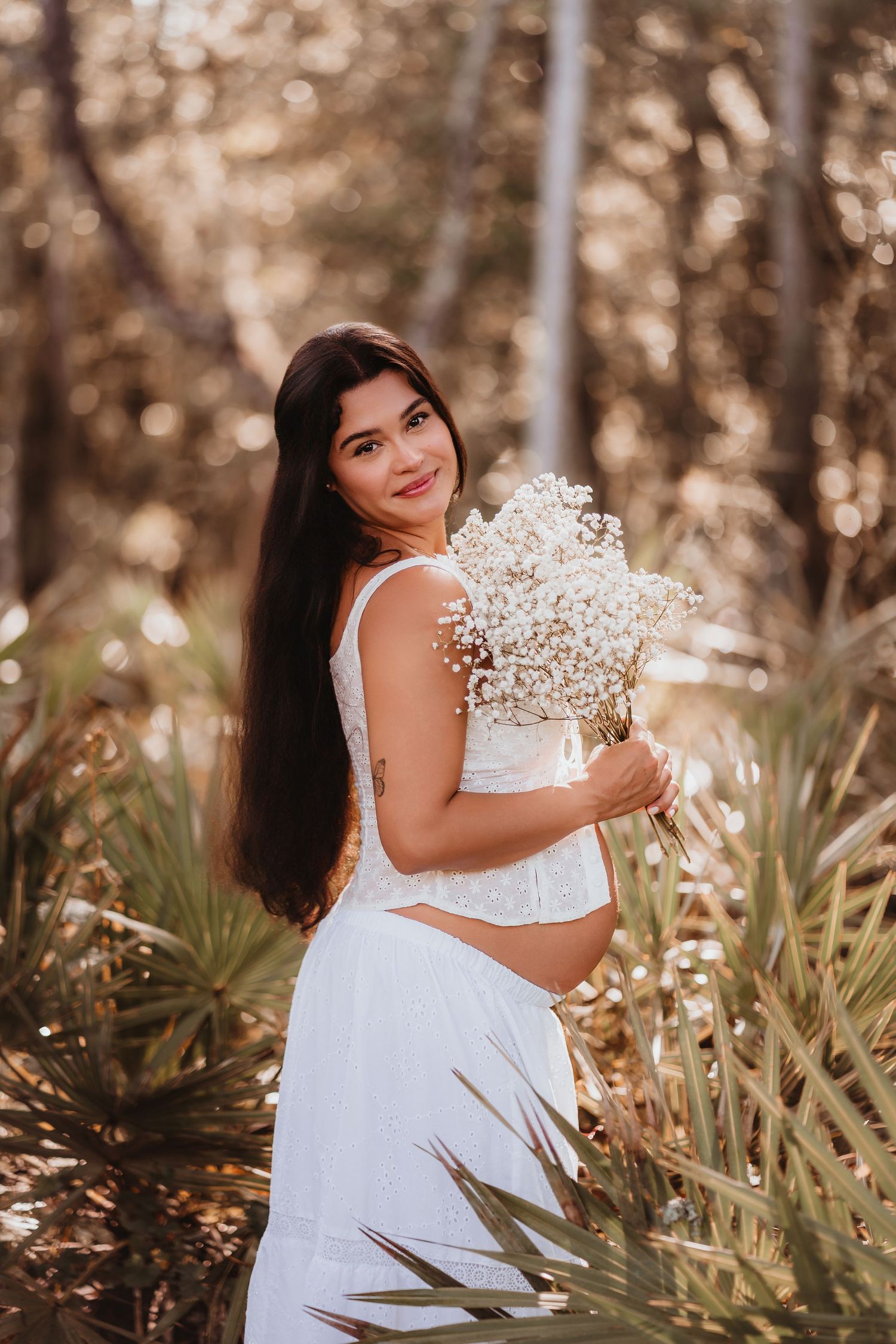 Pregnant woman in white dress holding baby's breath bouquet in a sunlit forest.