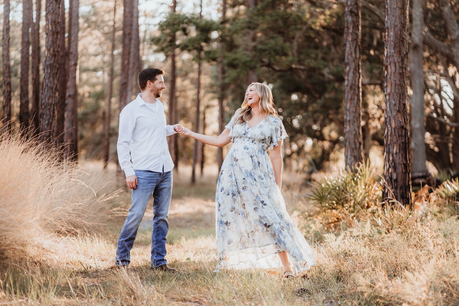 Happy expecting couple holding hands walking through a sunny pine forest path.