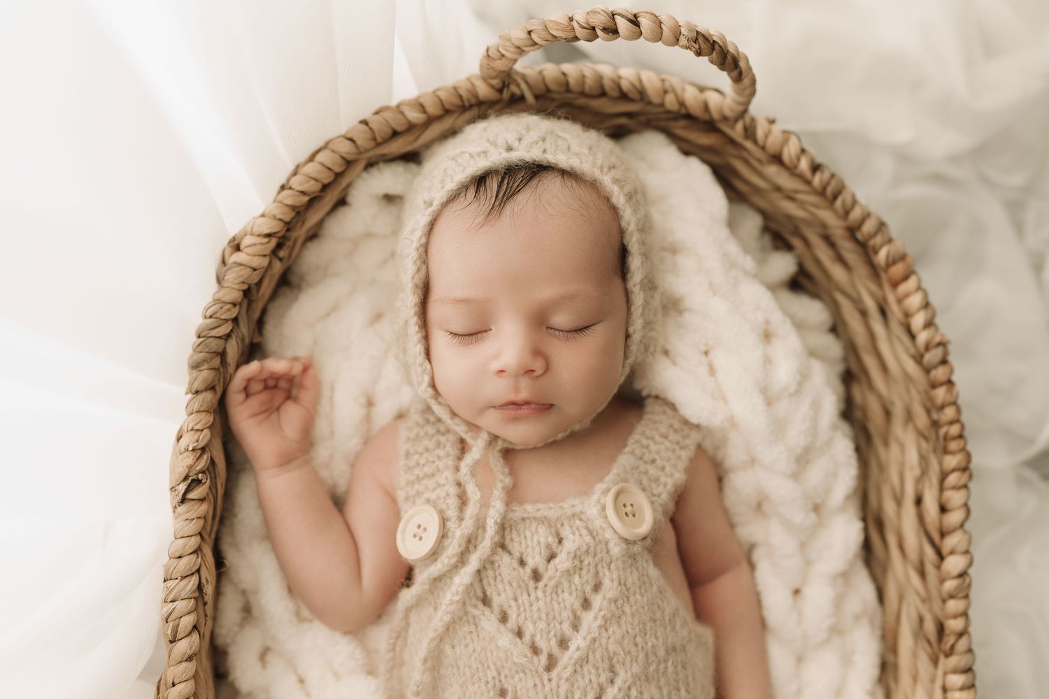 Sleeping newborn baby in knit romper and bonnet nestled in wicker basket with white chunky knit blanket.