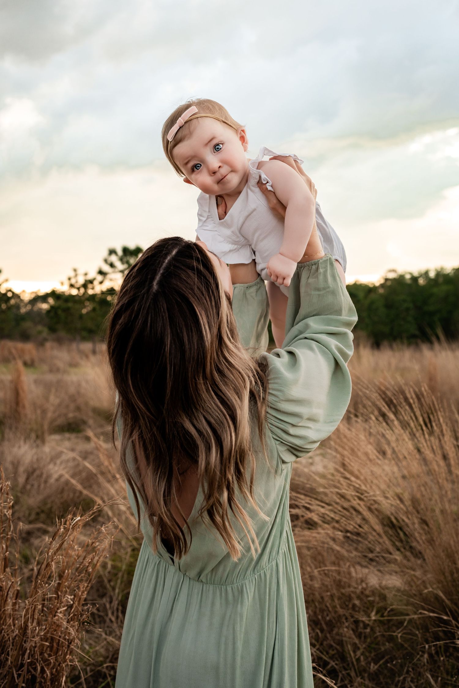 Mother in sage green dress lifting smiling baby girl overhead in a golden field at dusk.