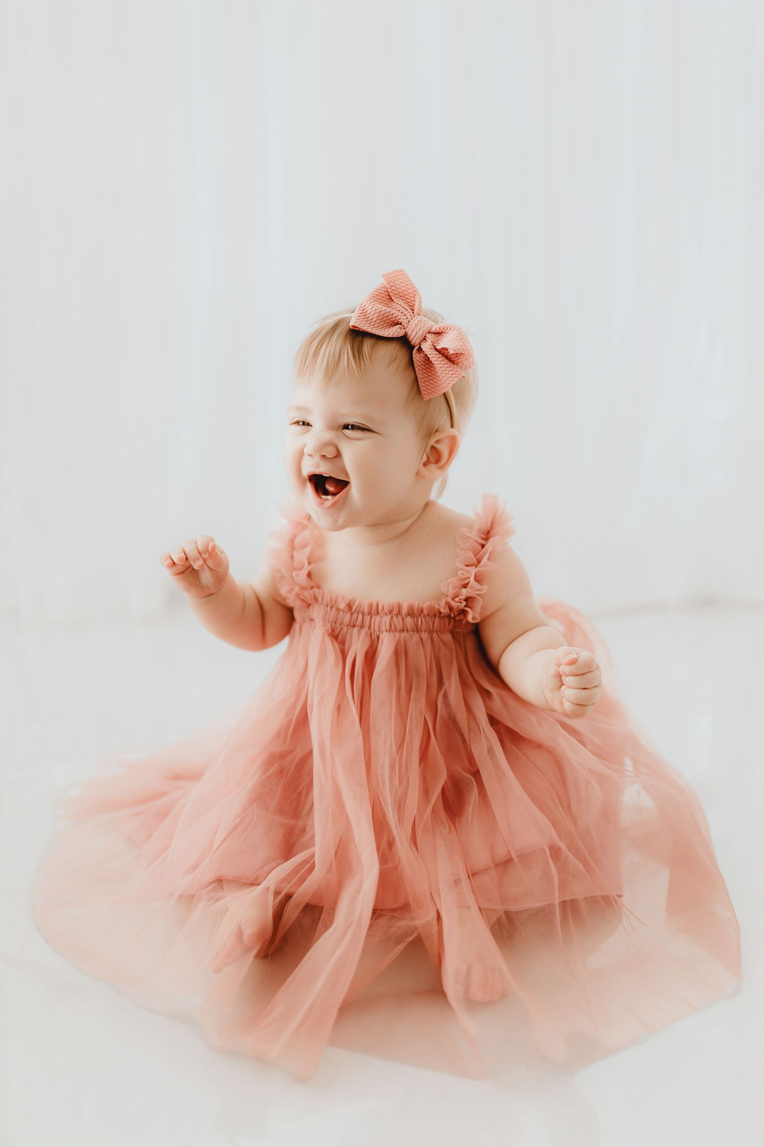 Laughing baby girl in pink tulle dress with bow headband reaching out joyfully.