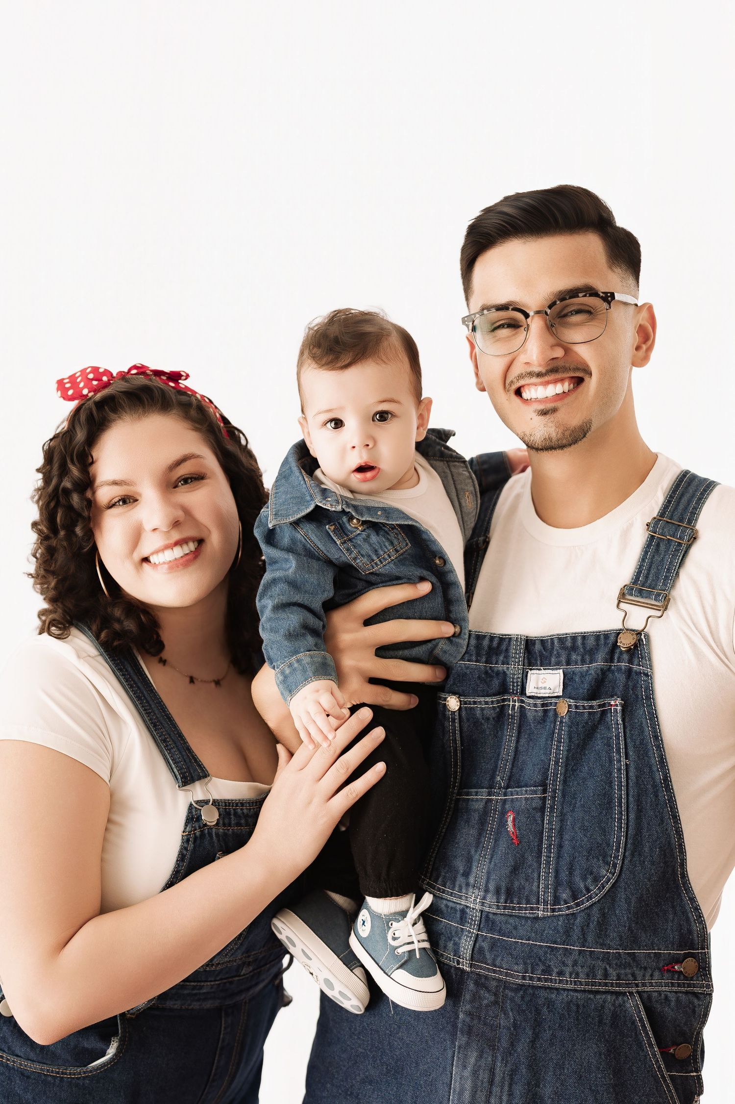Happy young family in matching denim outfits posing together with their cute baby on white background.
