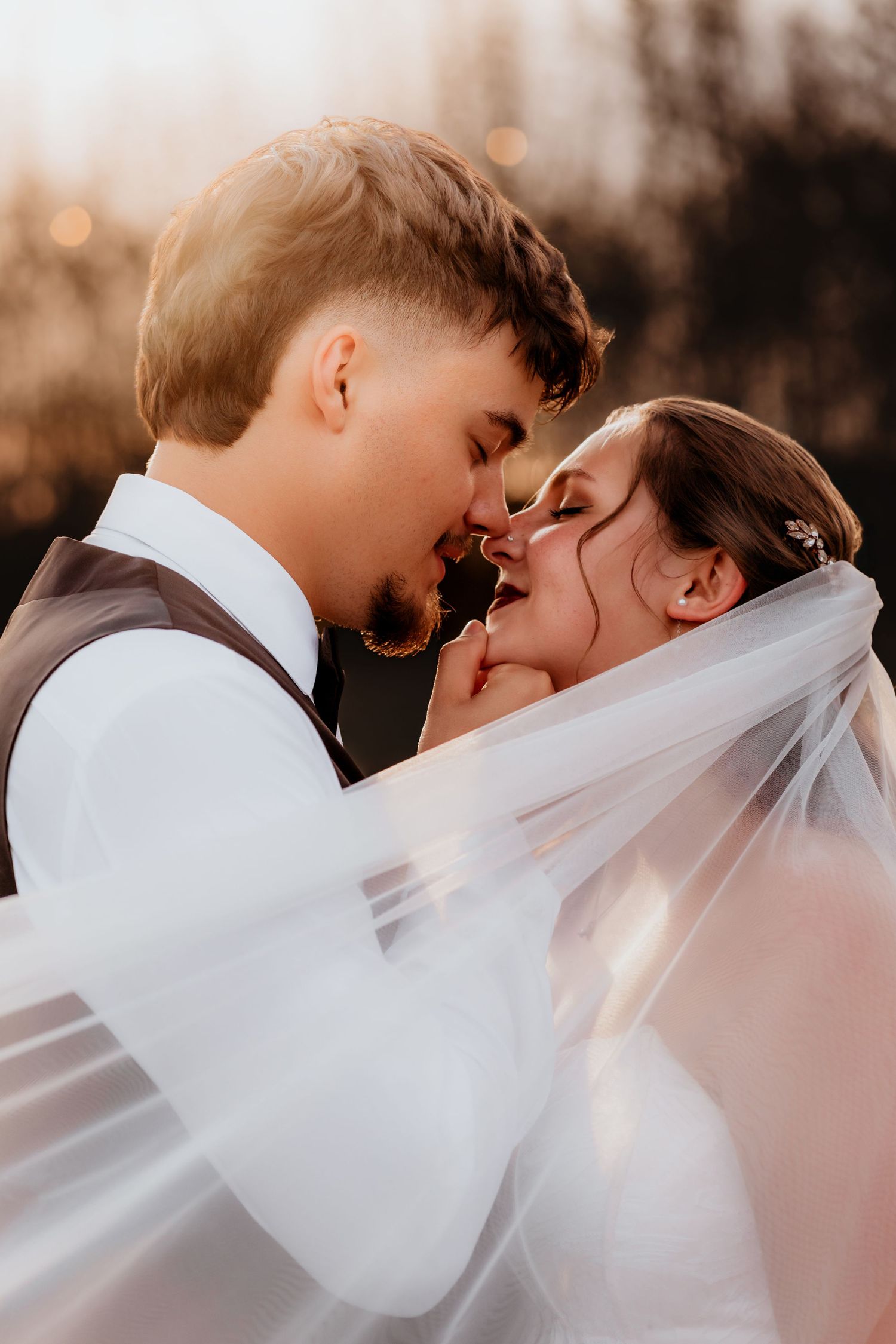 Bride and groom sharing a romantic almost-kiss under flowing white veil in golden sunset light.