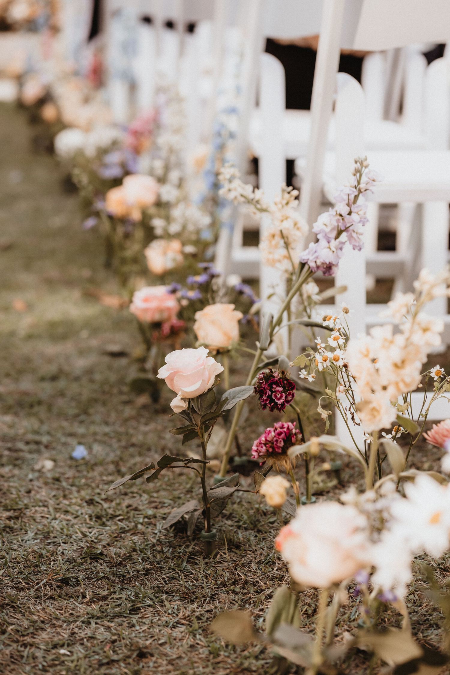 Elegant wedding aisle lined with pink roses and wildflowers beside white ceremony chairs outdoors.
