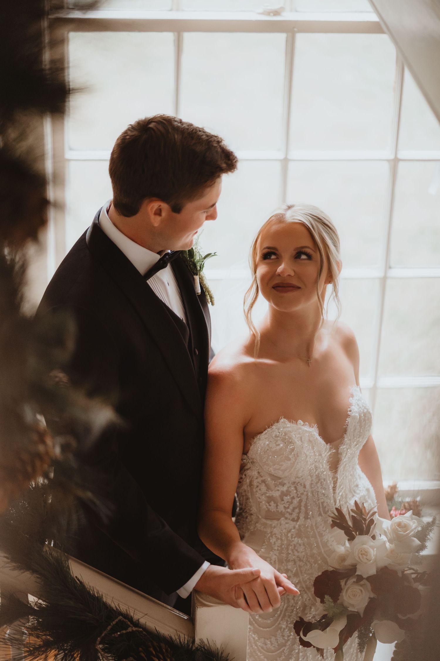 Bride in lace strapless gown and groom in tuxedo share a tender glance by a window.