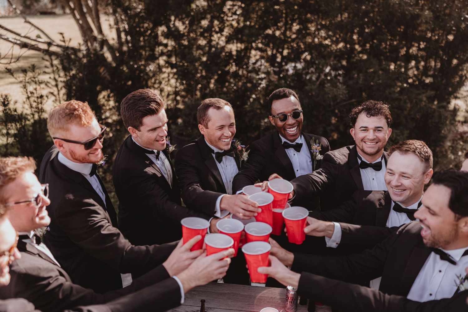 Wedding groomsmen in black tuxedos raising red solo cups in a celebratory toast outdoors.