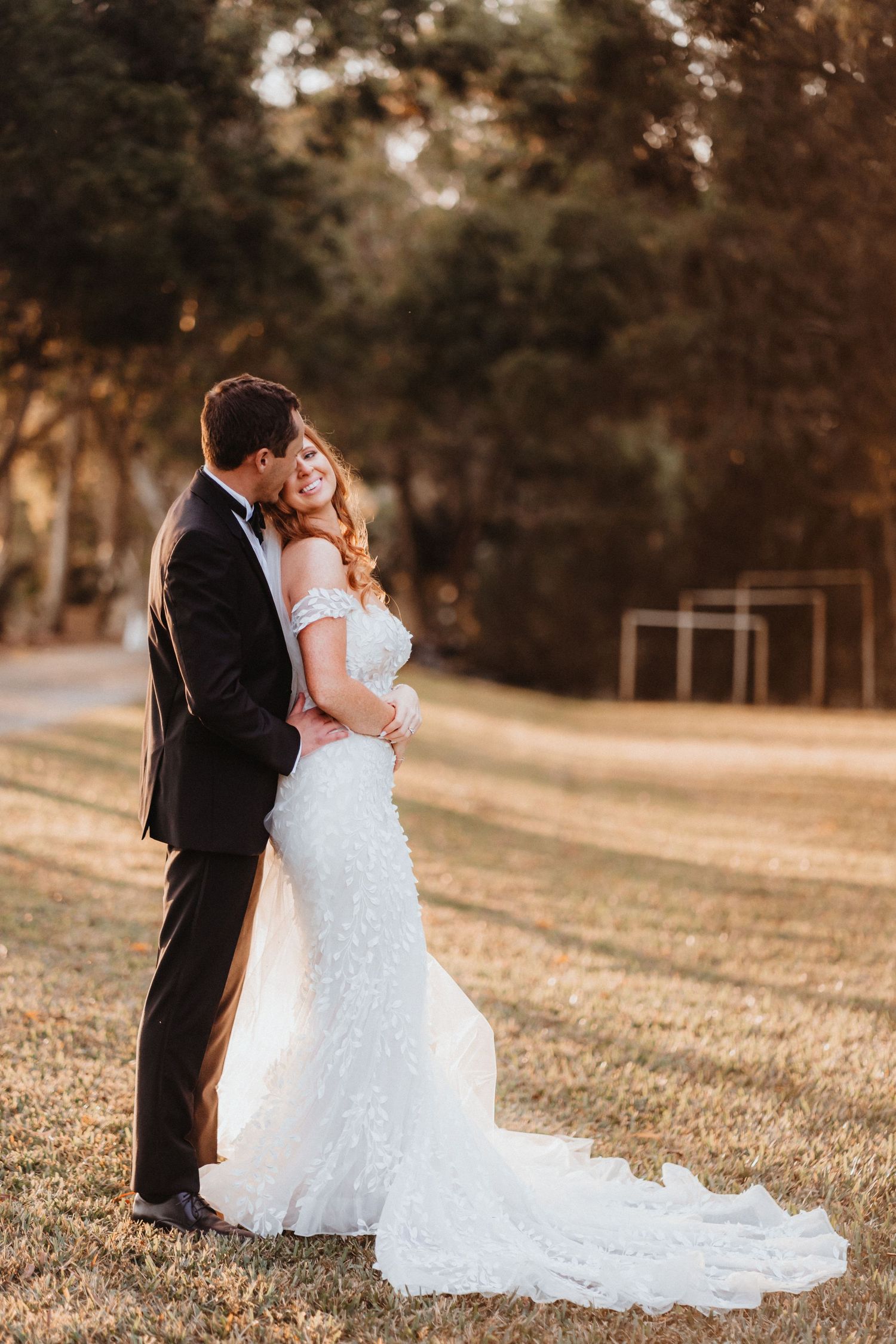 Groom in black tuxedo embraces bride in fitted lace gown with train during golden hour outdoor portrait.