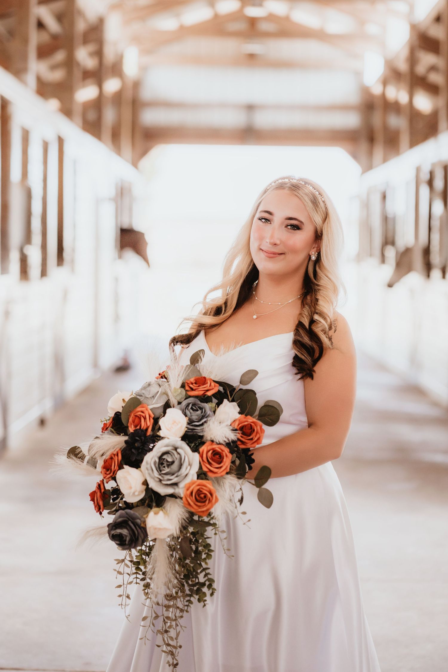 Smiling bride in a white strapless gown holds a cascading bouquet of orange, gray, and white roses in a barn.