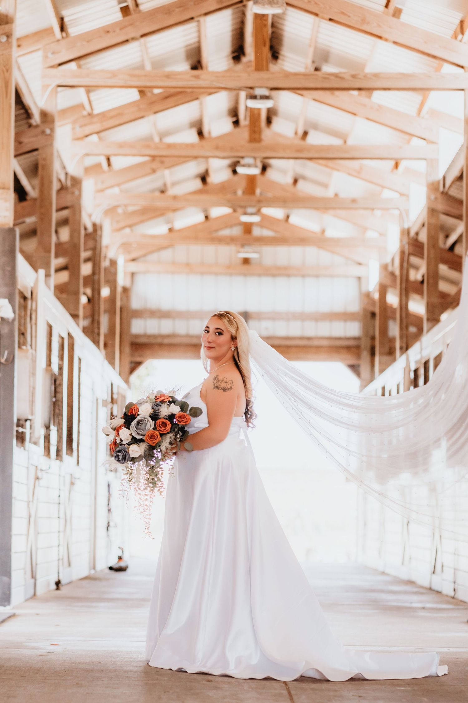 Bride in a white gown and flowing veil poses in a wooden horse stable holding a cascading floral bouquet.