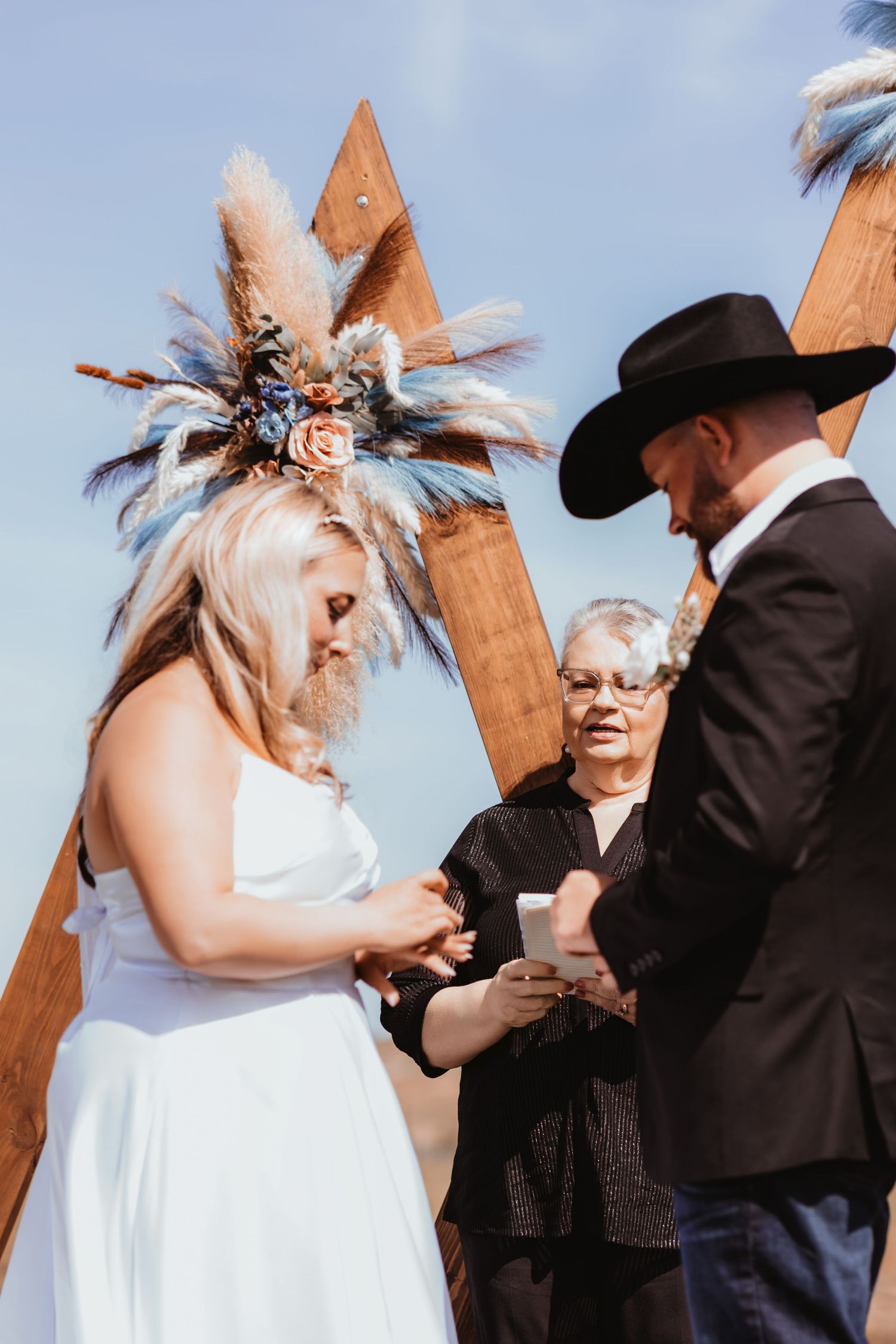 Bride places ring on groom's finger at rustic outdoor wedding ceremony with wooden triangular arch backdrop.
