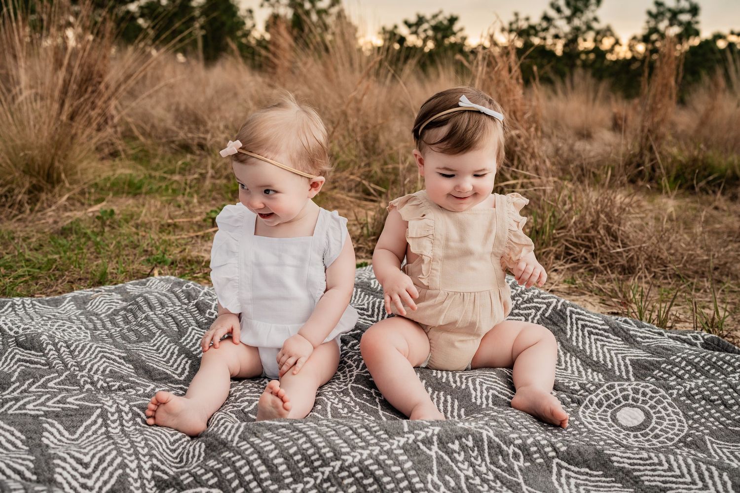 Two baby girls in white and beige rompers sitting on a patterned blanket in a golden field at sunset.