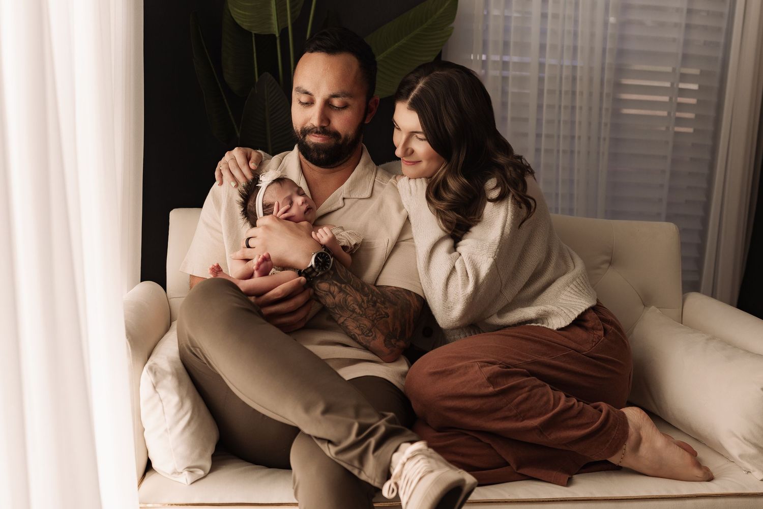 Young couple sitting on white sofa lovingly gazing at their newborn baby wrapped in pink blanket.