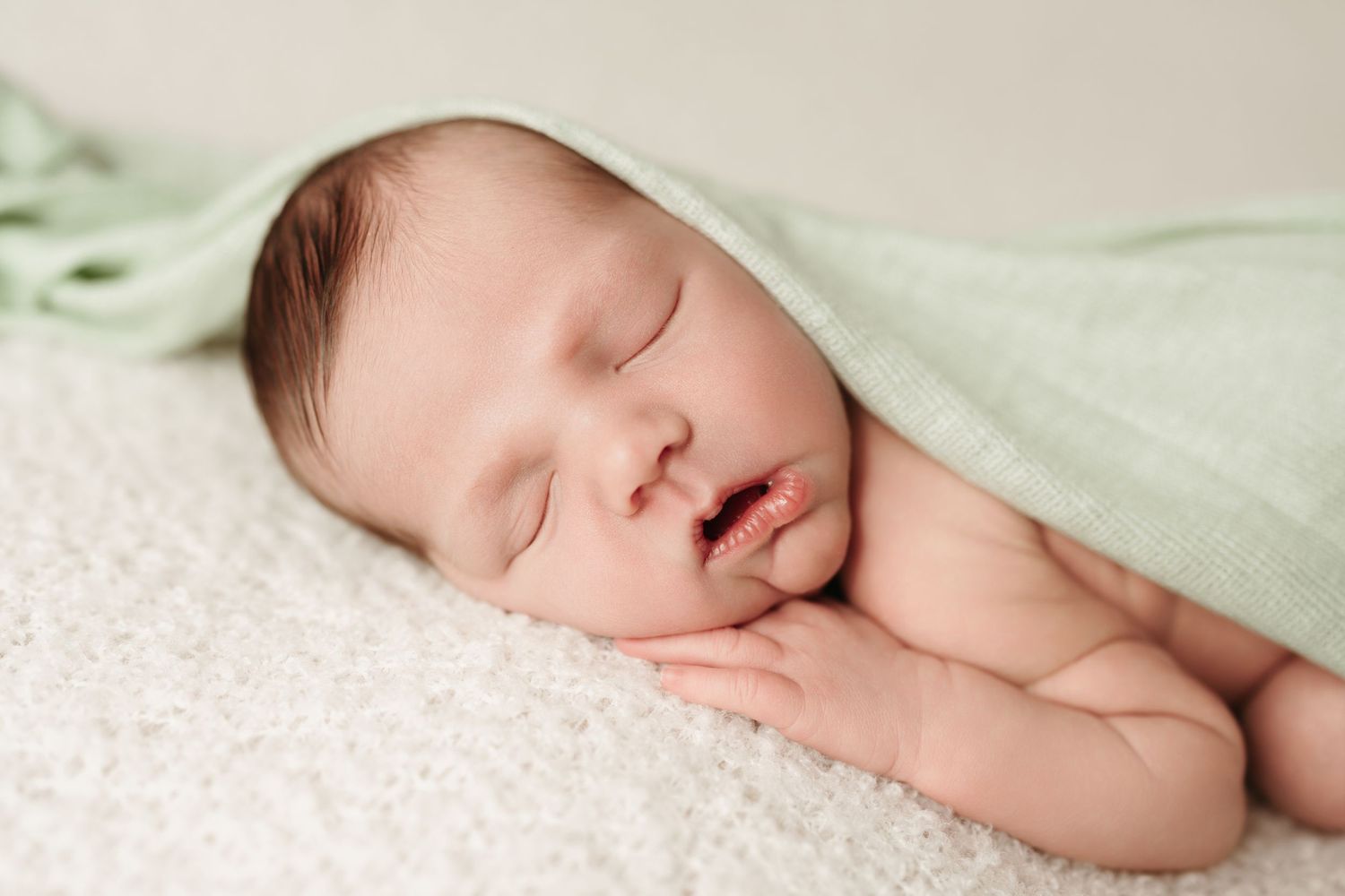 Serene newborn baby sleeping under light green wrap on white fluffy blanket.