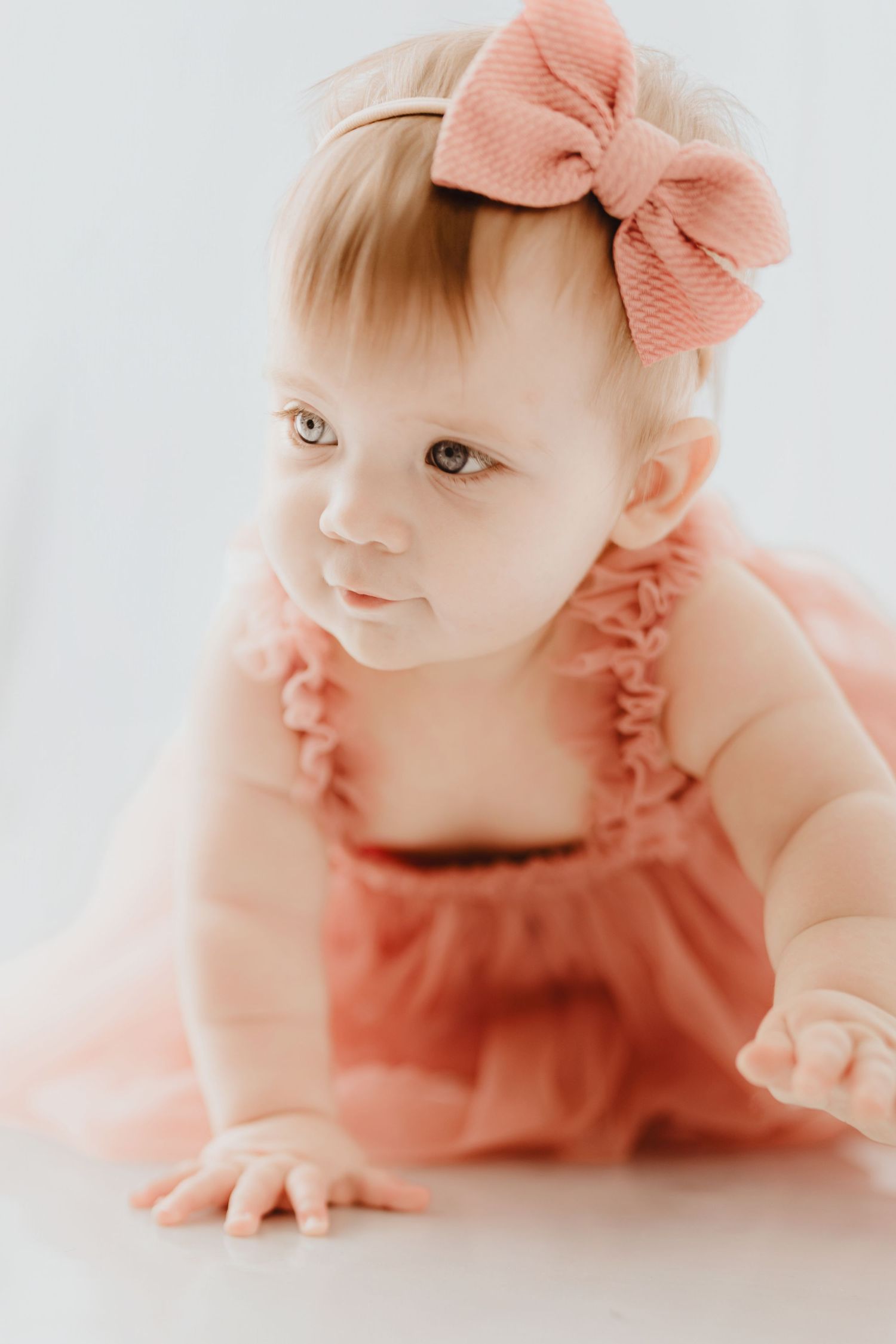 Curious baby girl in pink ruffle romper and bow headband crawling on white studio floor.
