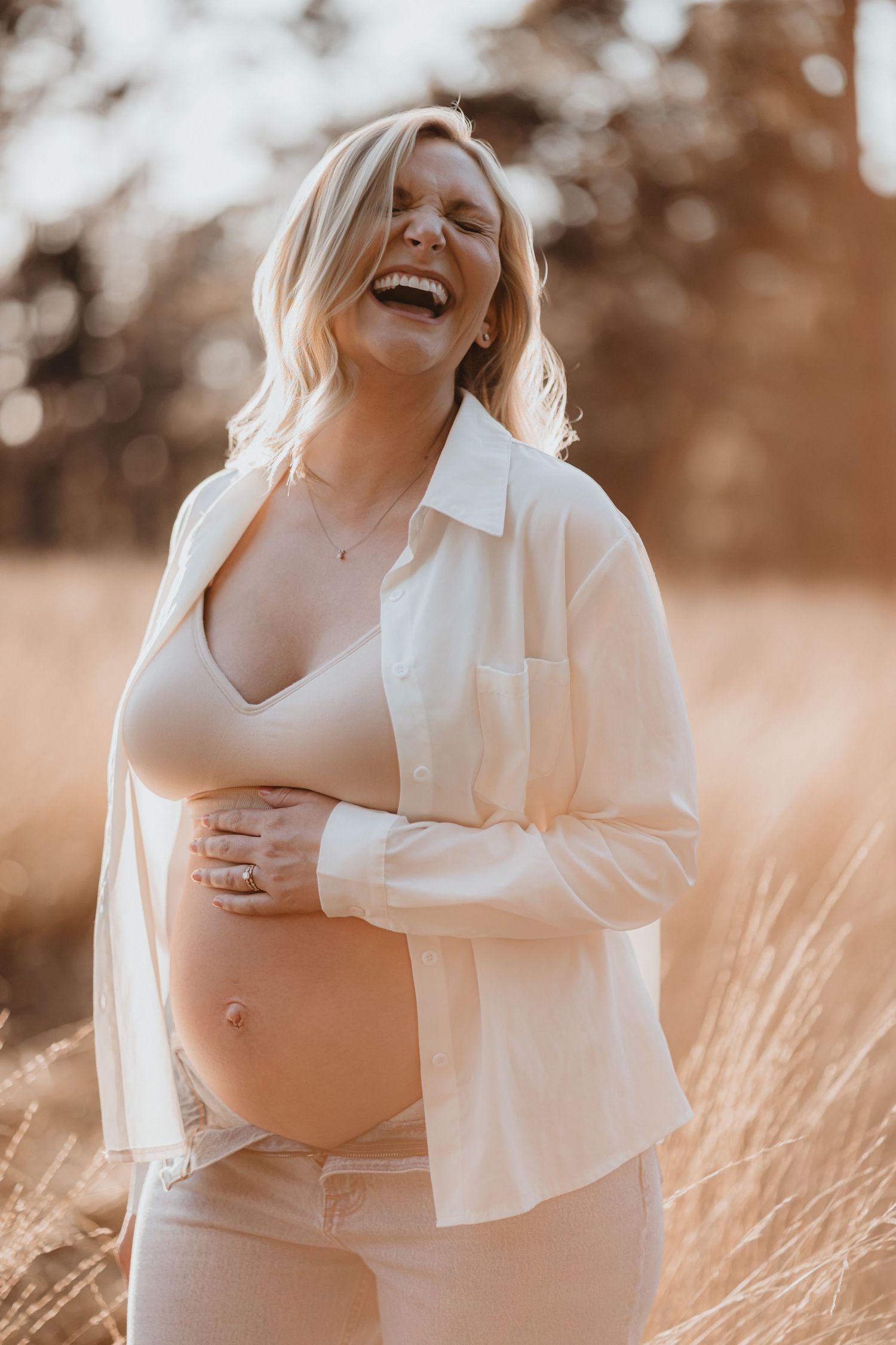 Laughing pregnant woman in open white shirt joyfully cradles bare baby bump in golden grass field.