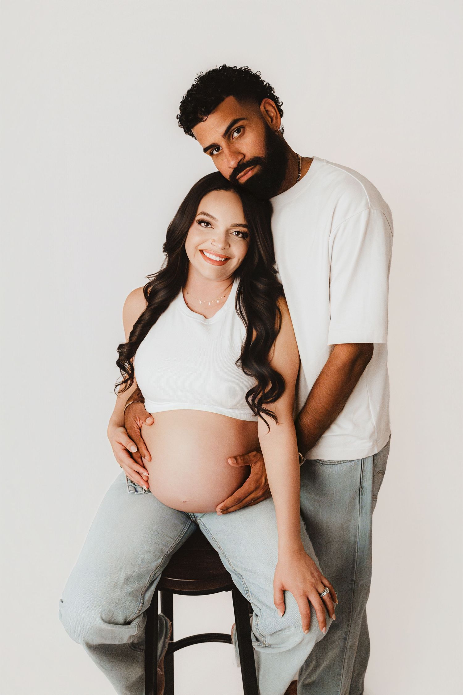 Happy pregnant couple in matching white outfits smiling at camera during maternity photo session.