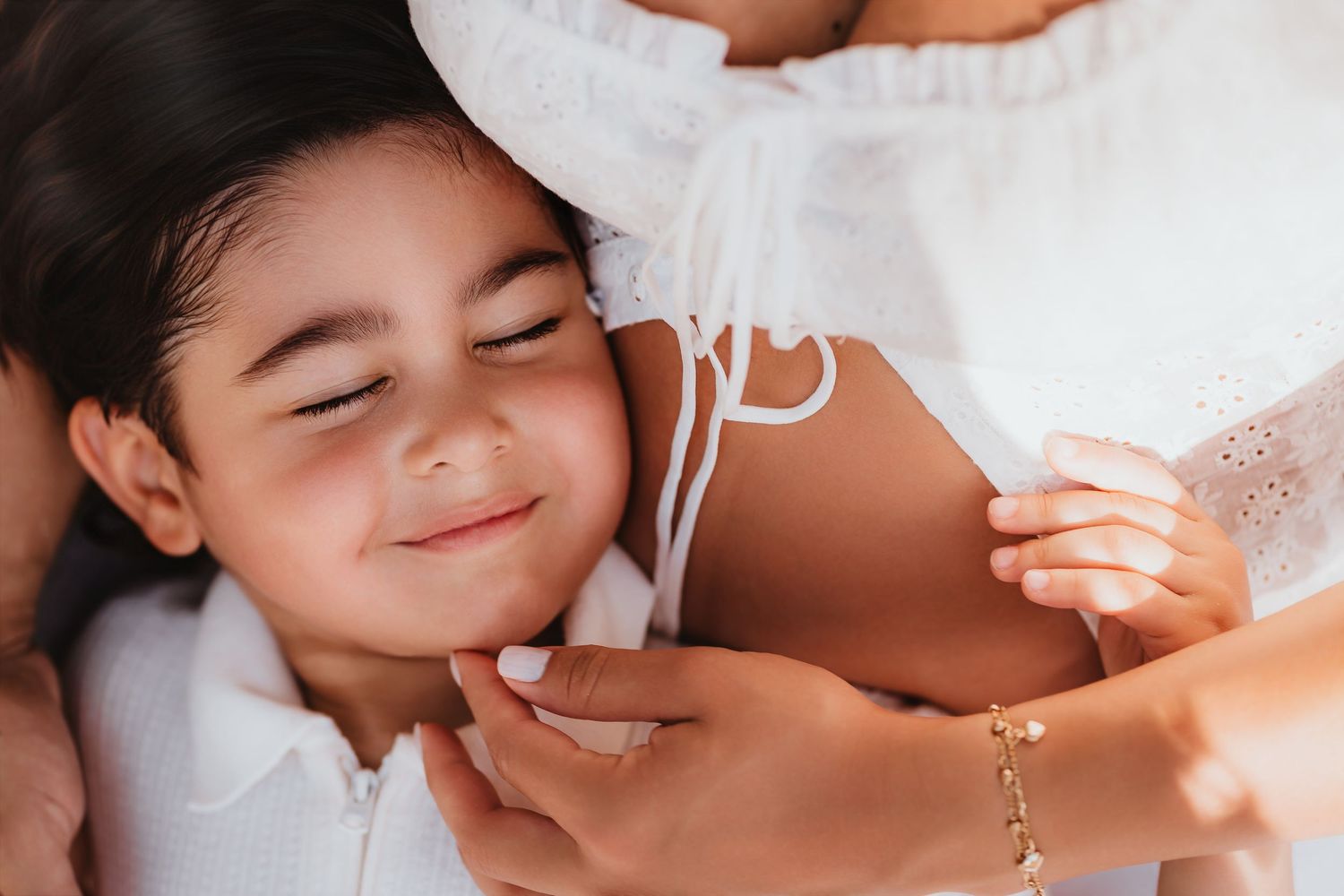 Young child smiling with eyes closed resting against pregnant mothers belly in white dress.