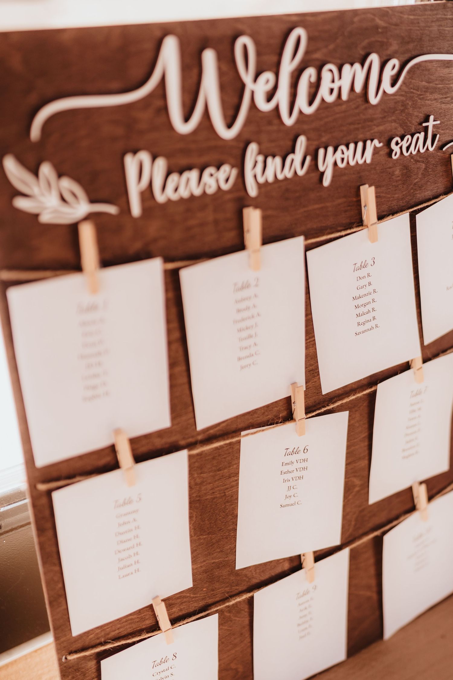Rustic wooden seating chart board with clothespin-attached table cards reads Welcome Please Find Your Seat.