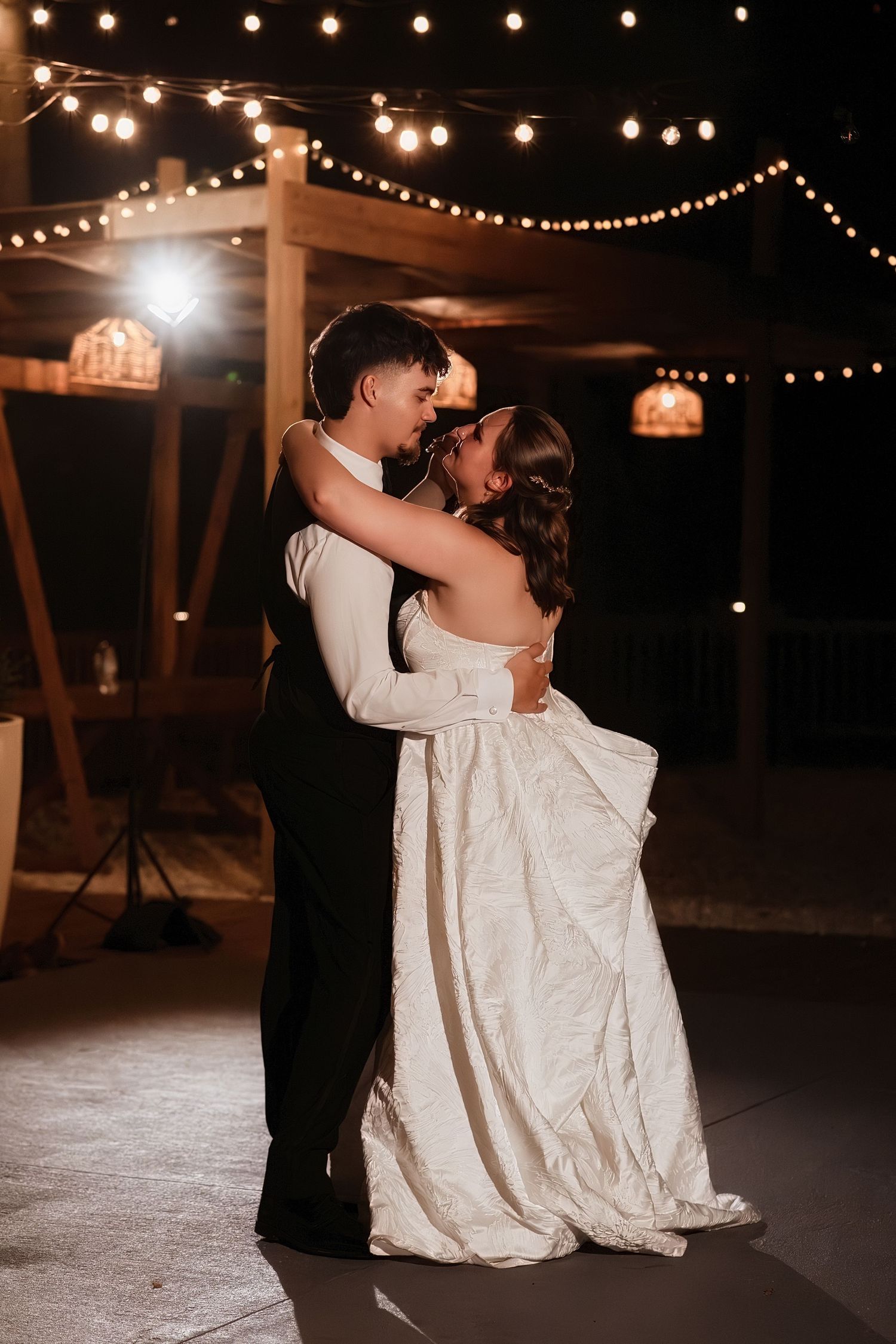 Bride and groom share romantic first dance under string lights at outdoor evening wedding reception.