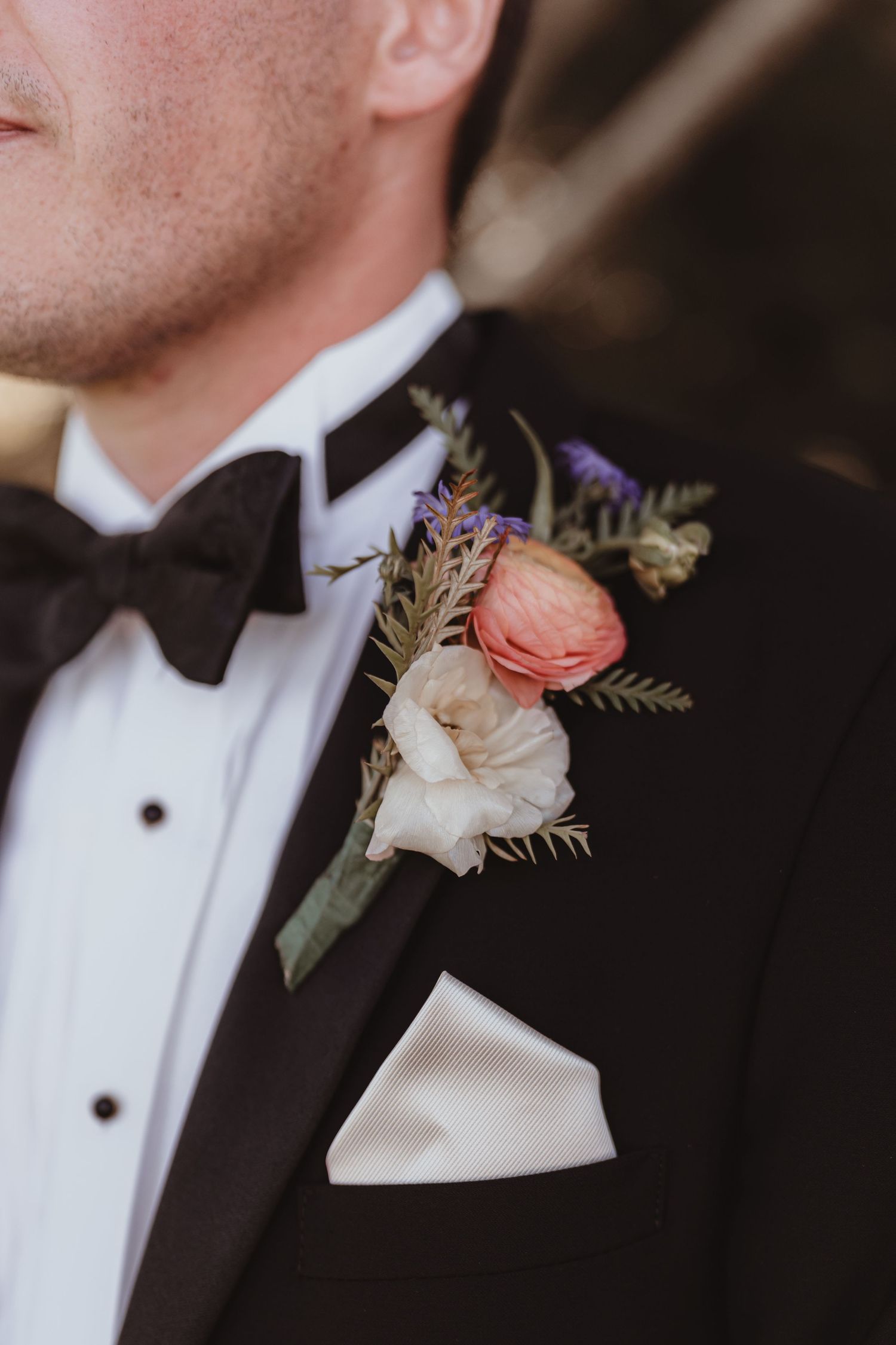 Close-up of a groom's boutonniere featuring pink ranunculus and white flowers on a black tuxedo lapel.