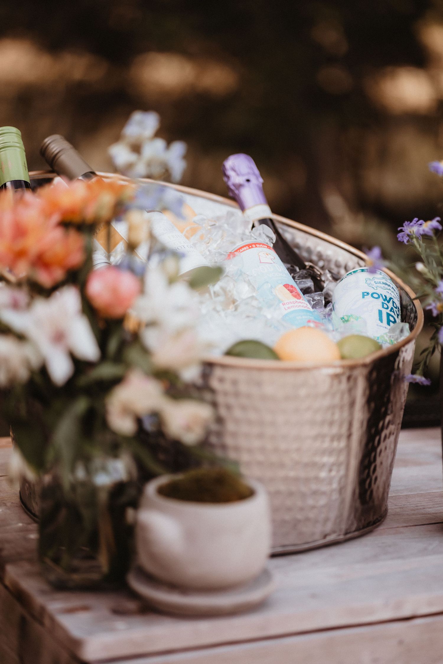Hammered metal ice bucket with champagne and drinks surrounded by fresh flowers at a wedding.