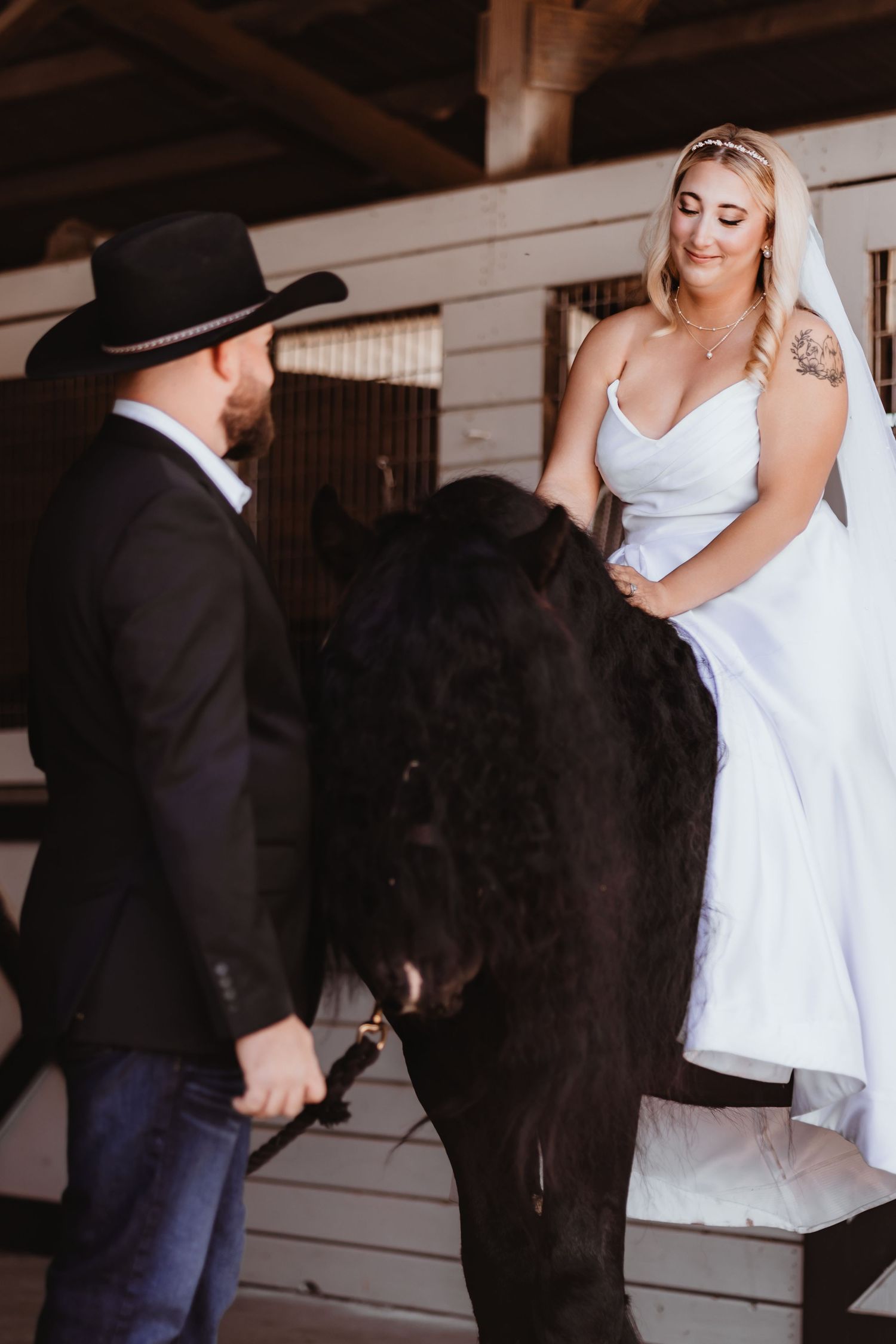 Groom in cowboy hat helps bride in white dress mount a black horse in barn.