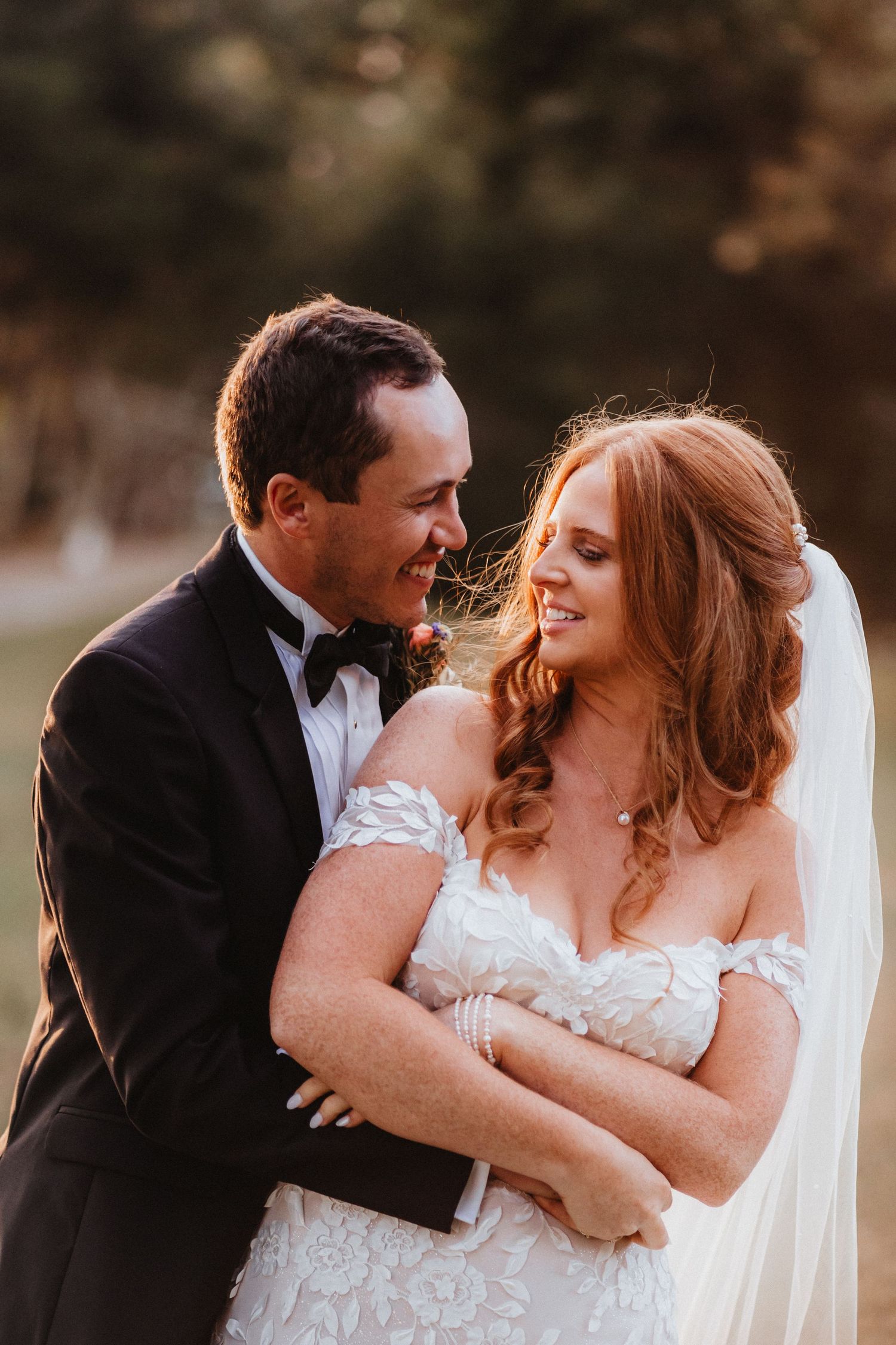 Groom tenderly embraces smiling bride at sunset, both looking at each other, bride in lace gown with flowing veil.