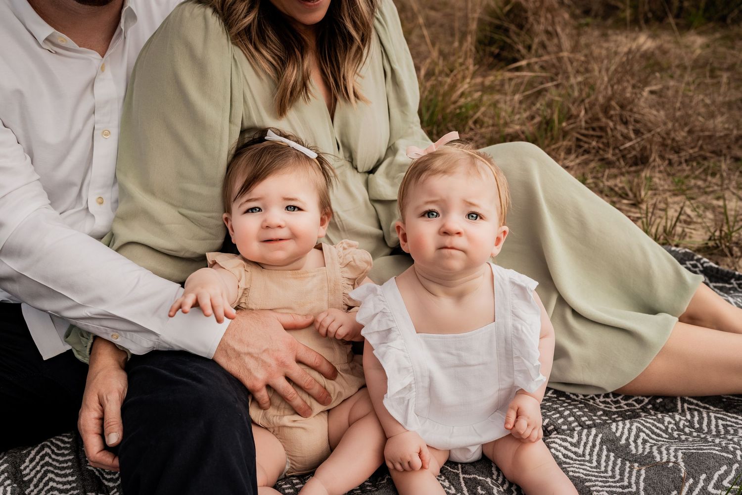 Two baby girls sitting on a blanket outdoors, one in a beige romper and one in white, with parents nearby.