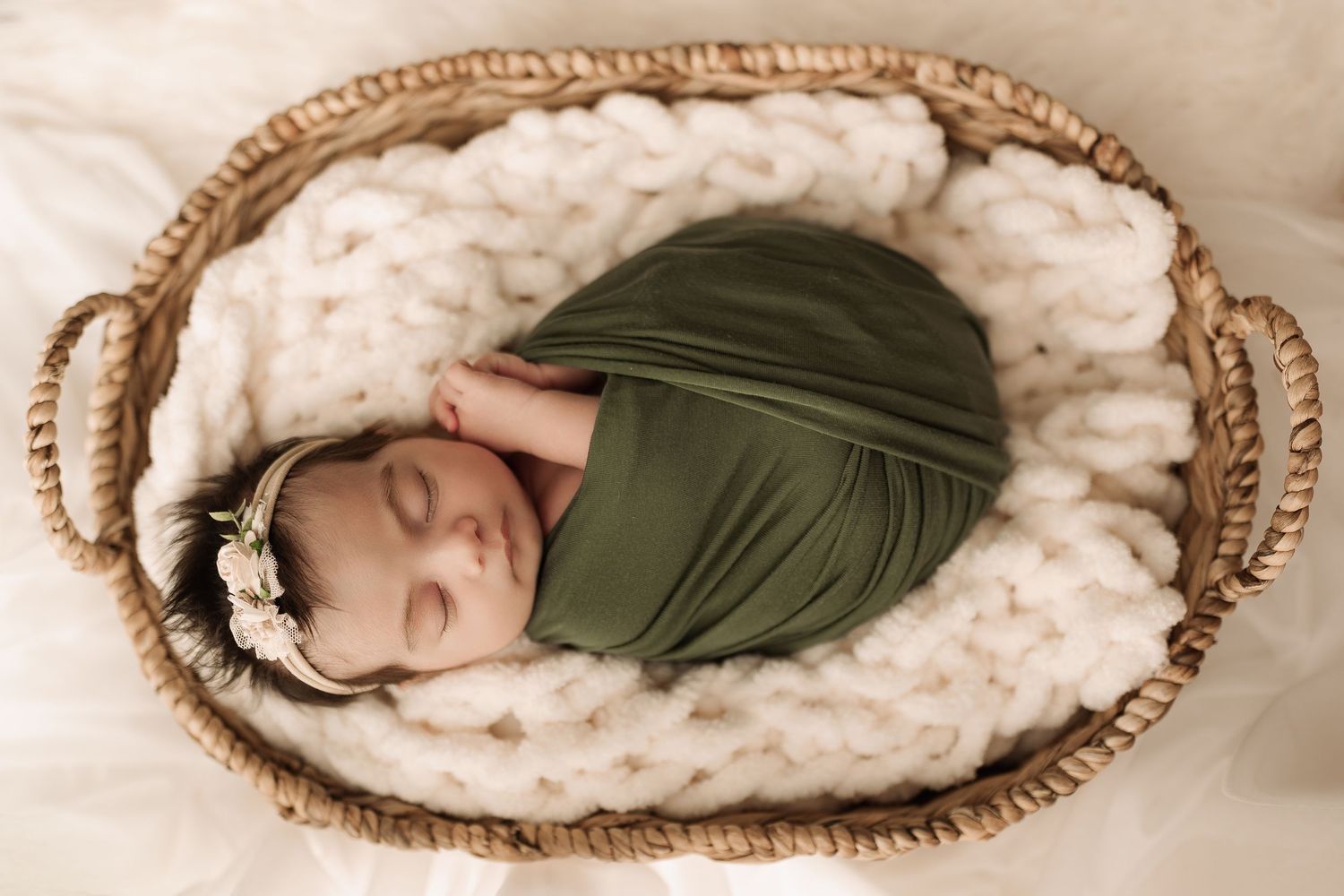 Peaceful newborn baby in olive green wrap and floral headband sleeping in wicker basket.