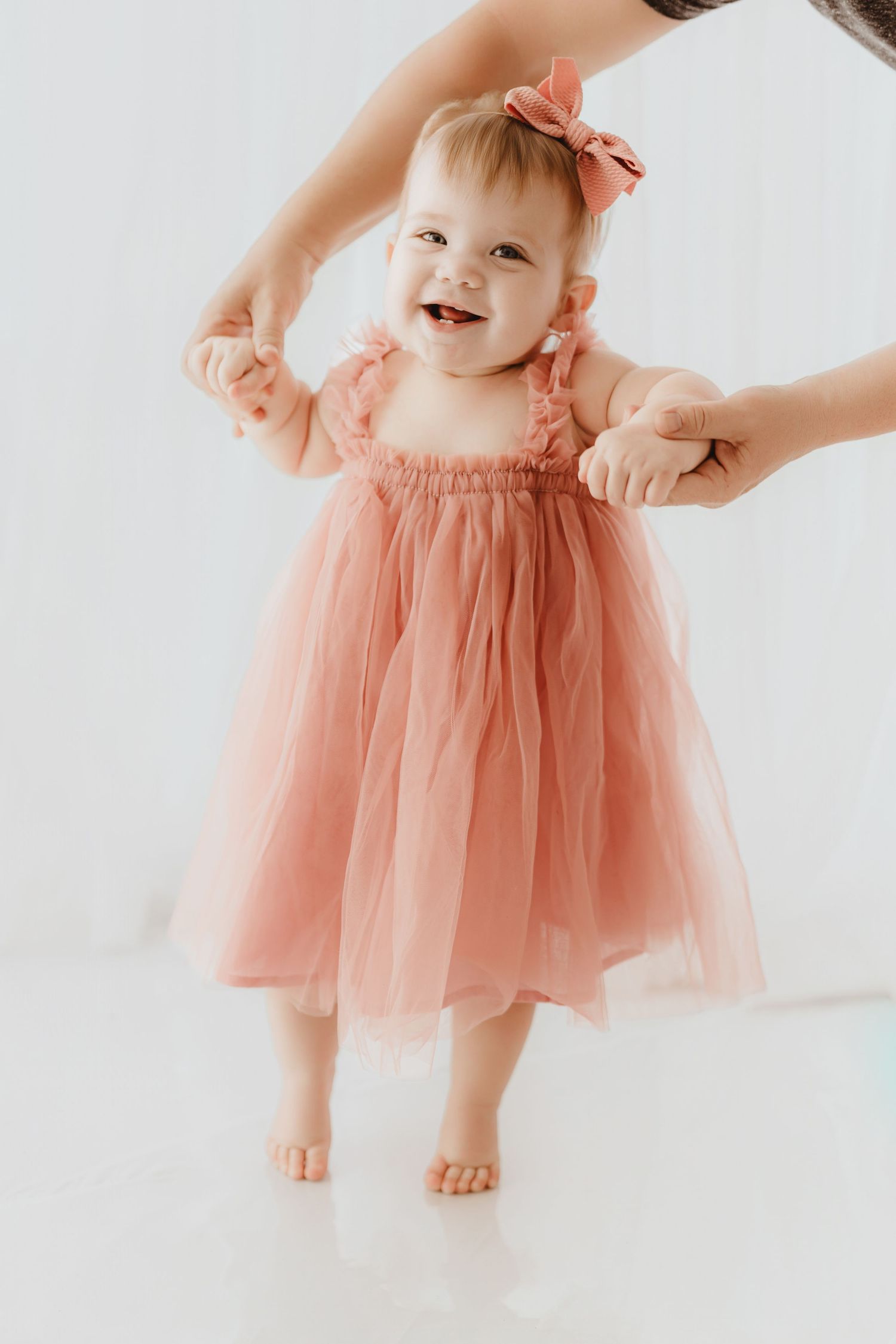 Laughing baby girl in dusty rose tulle dress practices walking with help.