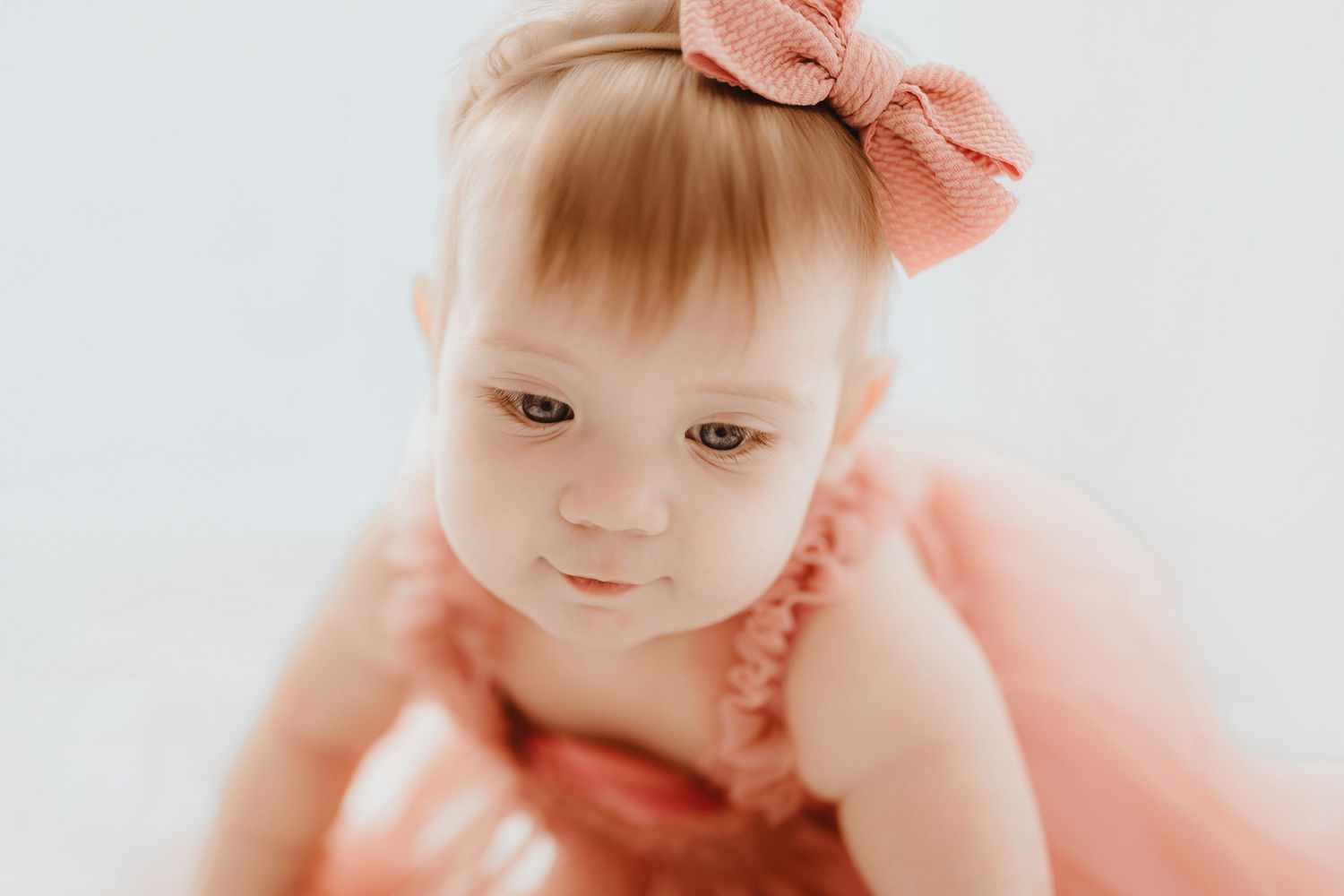 Sweet baby girl in pink ruffle dress with bow headband looking down softly.