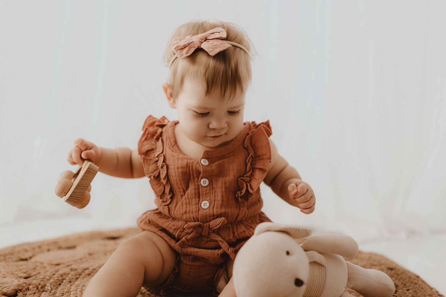 Baby girl in rust romper playing with wooden toy beside stuffed bunny on rug.