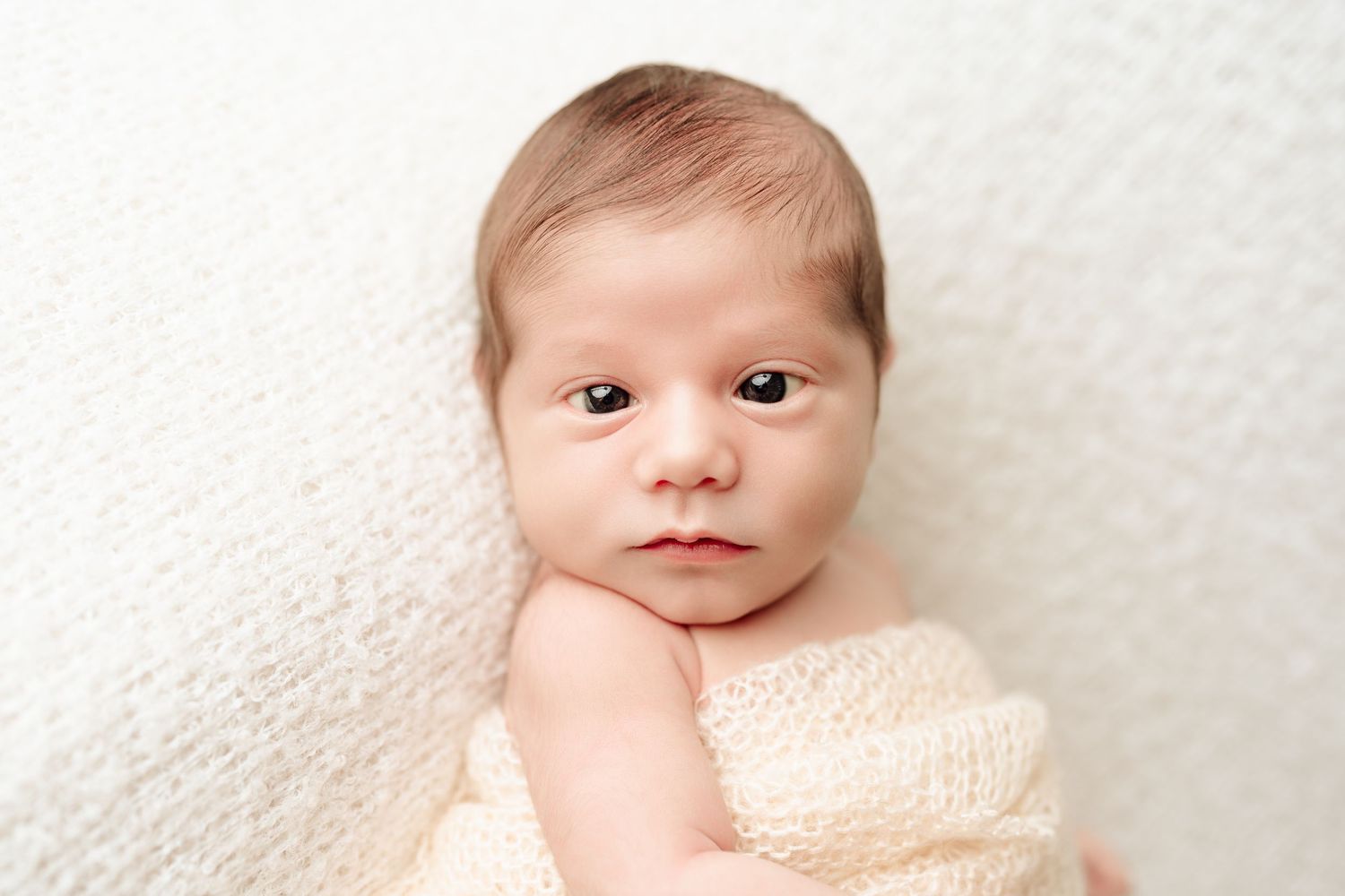 Newborn baby wrapped in cream knit blanket lying on white textured background.