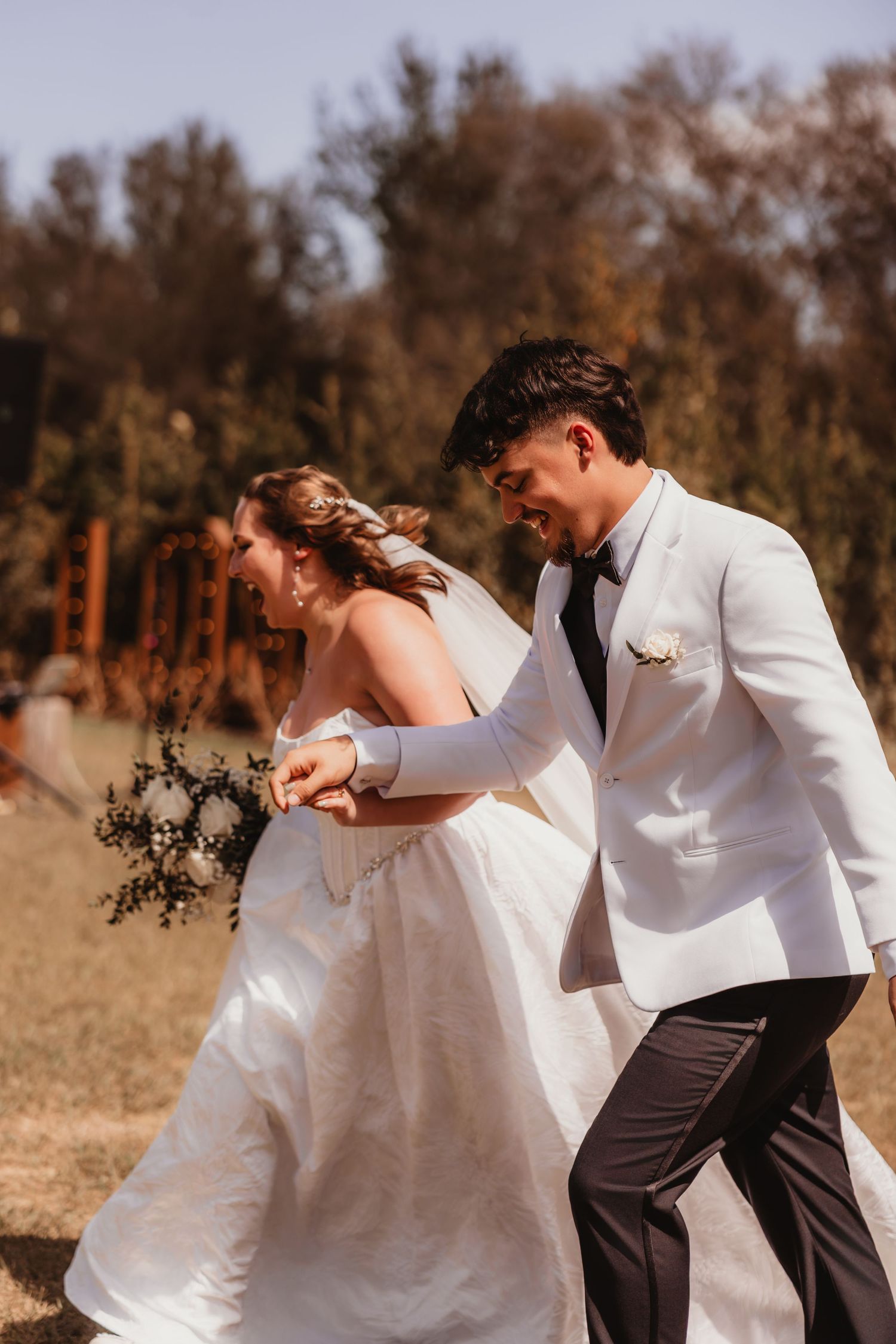 Bride in white strapless gown and groom in white tuxedo jacket walking together in sunny outdoor setting.