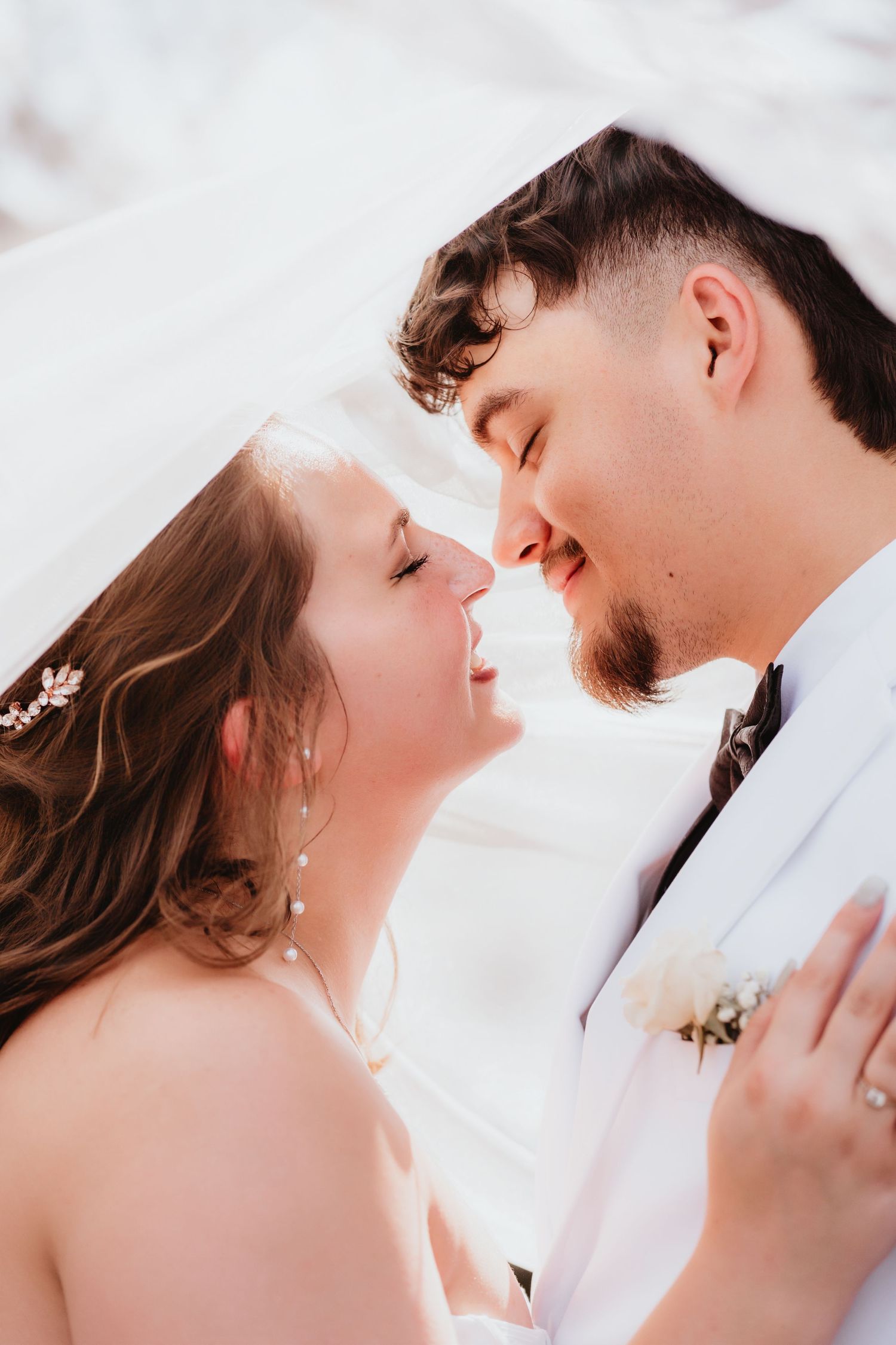 Tender moment of smiling bride and groom leaning in for a kiss under flowing white veil.